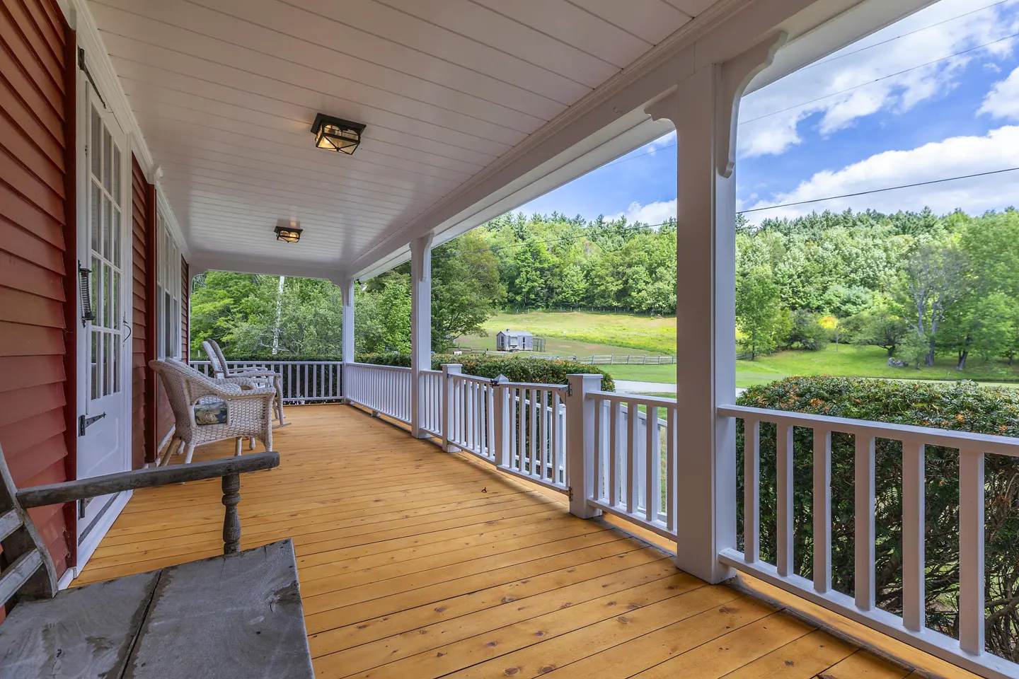 Covered porch with wood floor, white railing, and wicker chairs. A green field and trees are visible in the background.