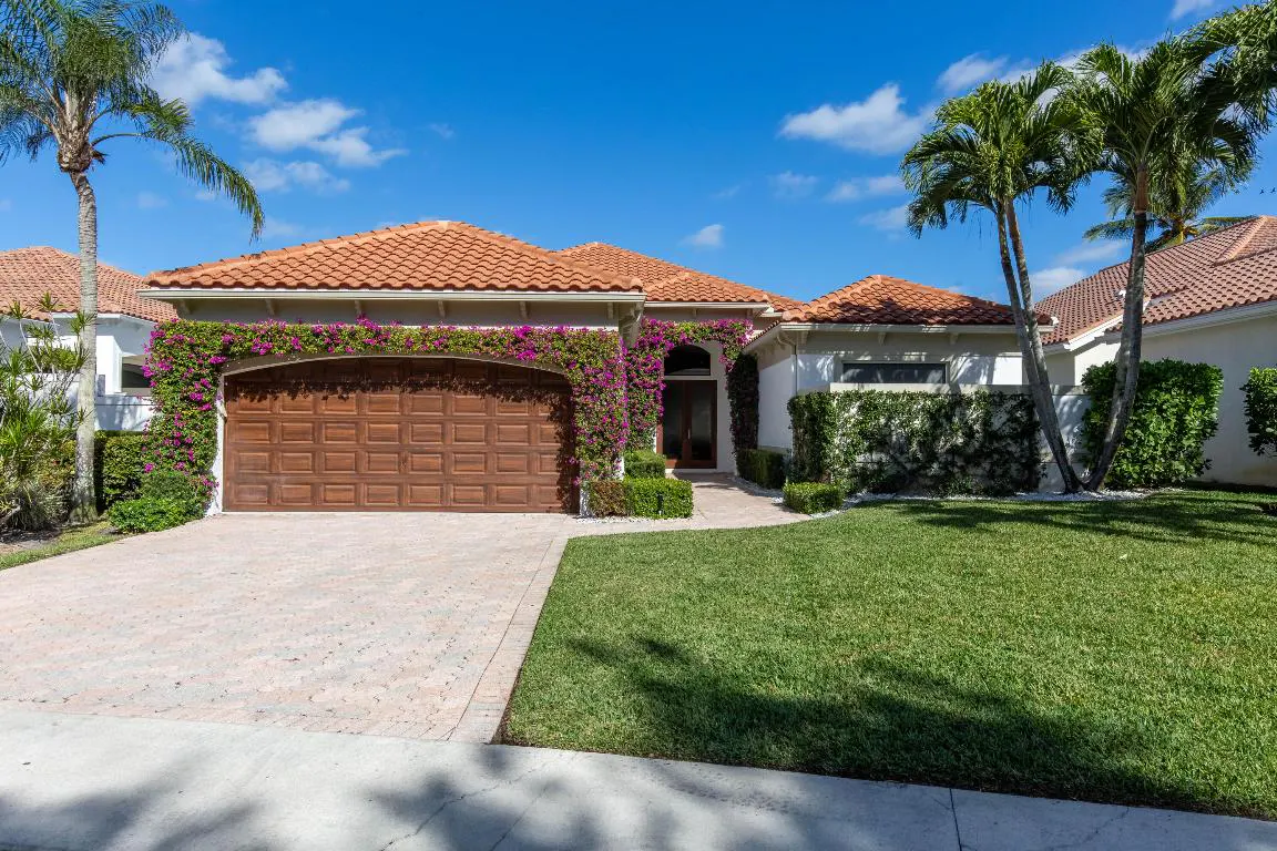 A single-story house with a brown garage door and a red tile roof, framed by palm trees and a blue sky.
