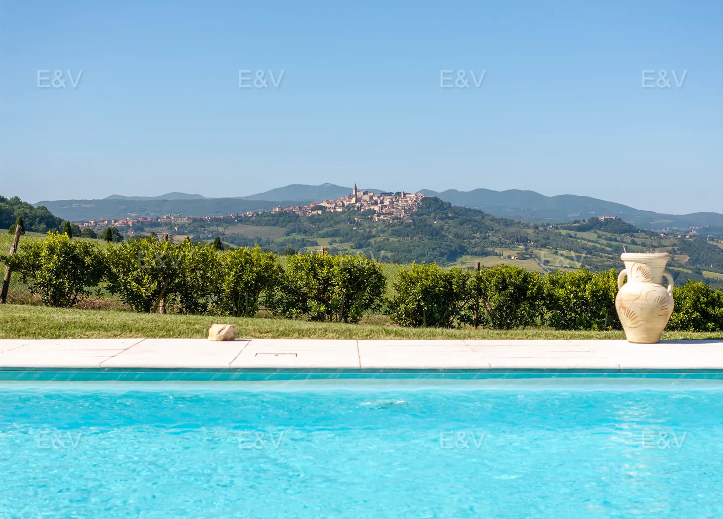 A turquoise pool with a white border overlooks green hills and a distant town under a clear blue sky. A large, decorative vase sits poolside.