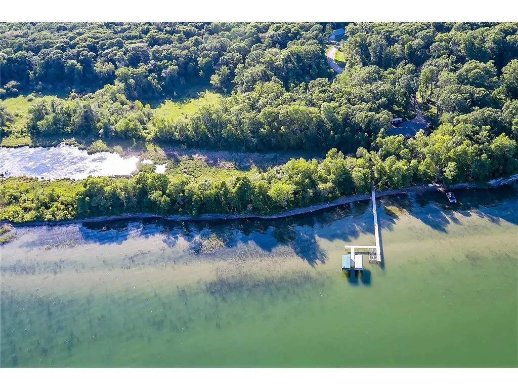 Aerial view of a lakefront property with a long white dock and a green boathouse. The shoreline is lined with lush green trees.