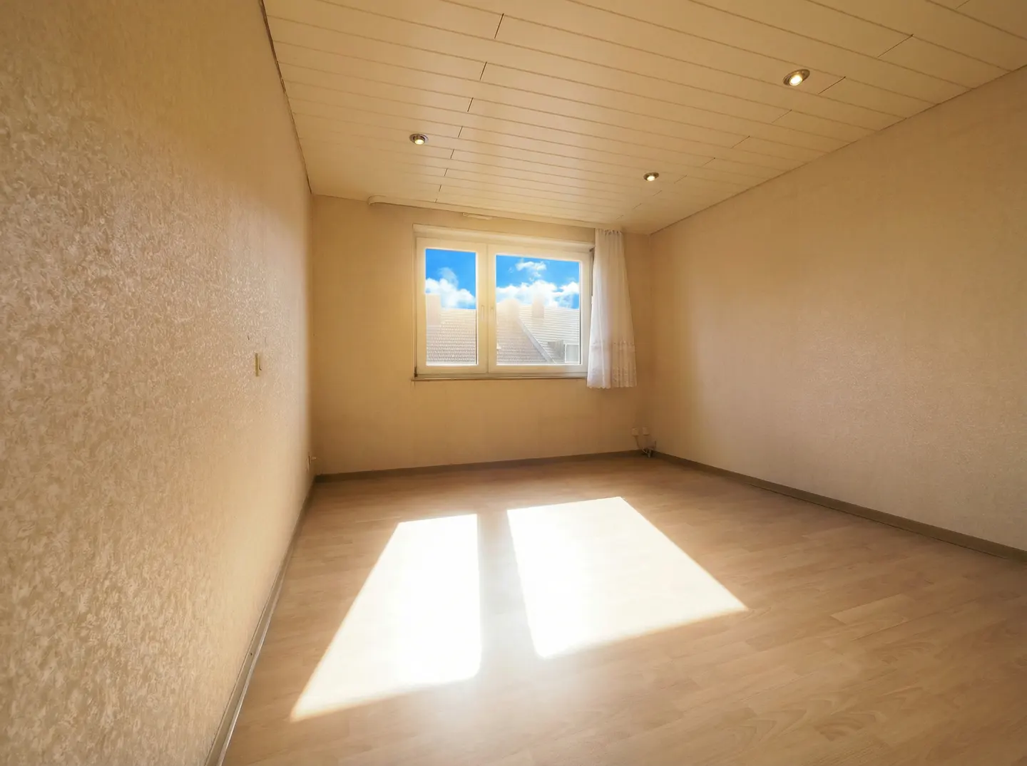 Empty room with beige walls, wood floor, and a window showing a blue sky. Sunlight streams in, creating bright patches on the floor.