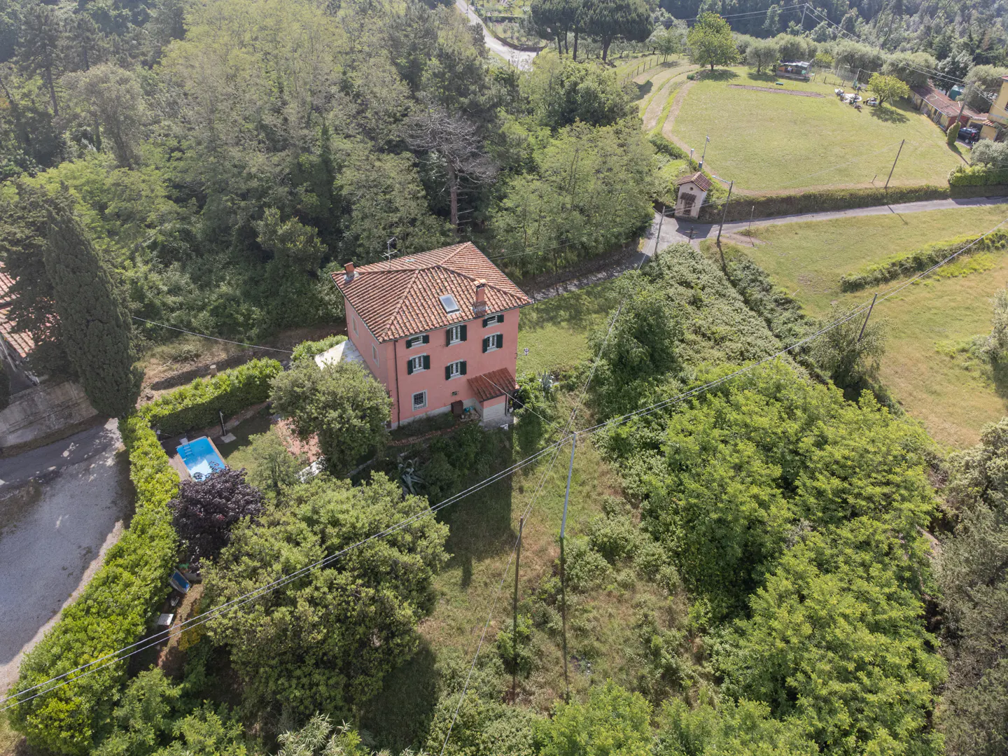 Aerial view of a pink three-story house with a tiled roof, a small swimming pool, and surrounding trees and fields.