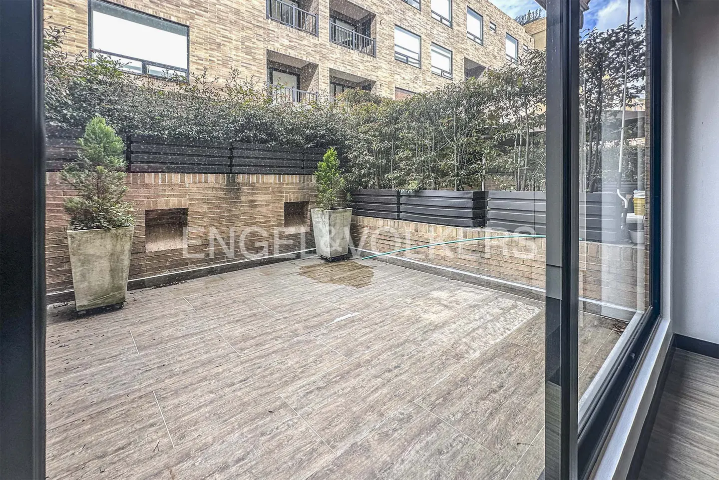 A patio with light brown tiles, brick walls, and greenery, viewed through a sliding glass door.