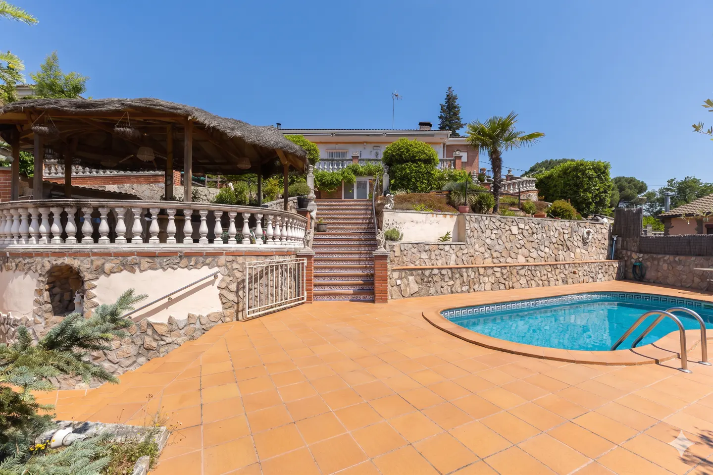 Outdoor view of a luxury home with a pool, stone walls, and a thatched-roof gazebo on a sunny day.