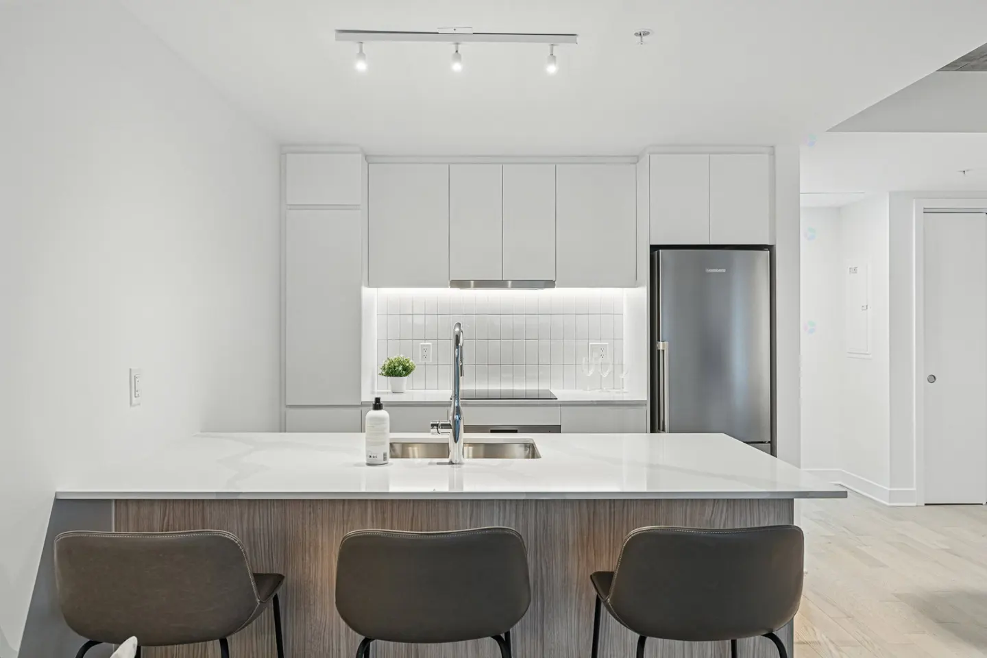 Bright, modern kitchen with white cabinets, stainless steel fridge, and a marble countertop island with three gray bar stools.