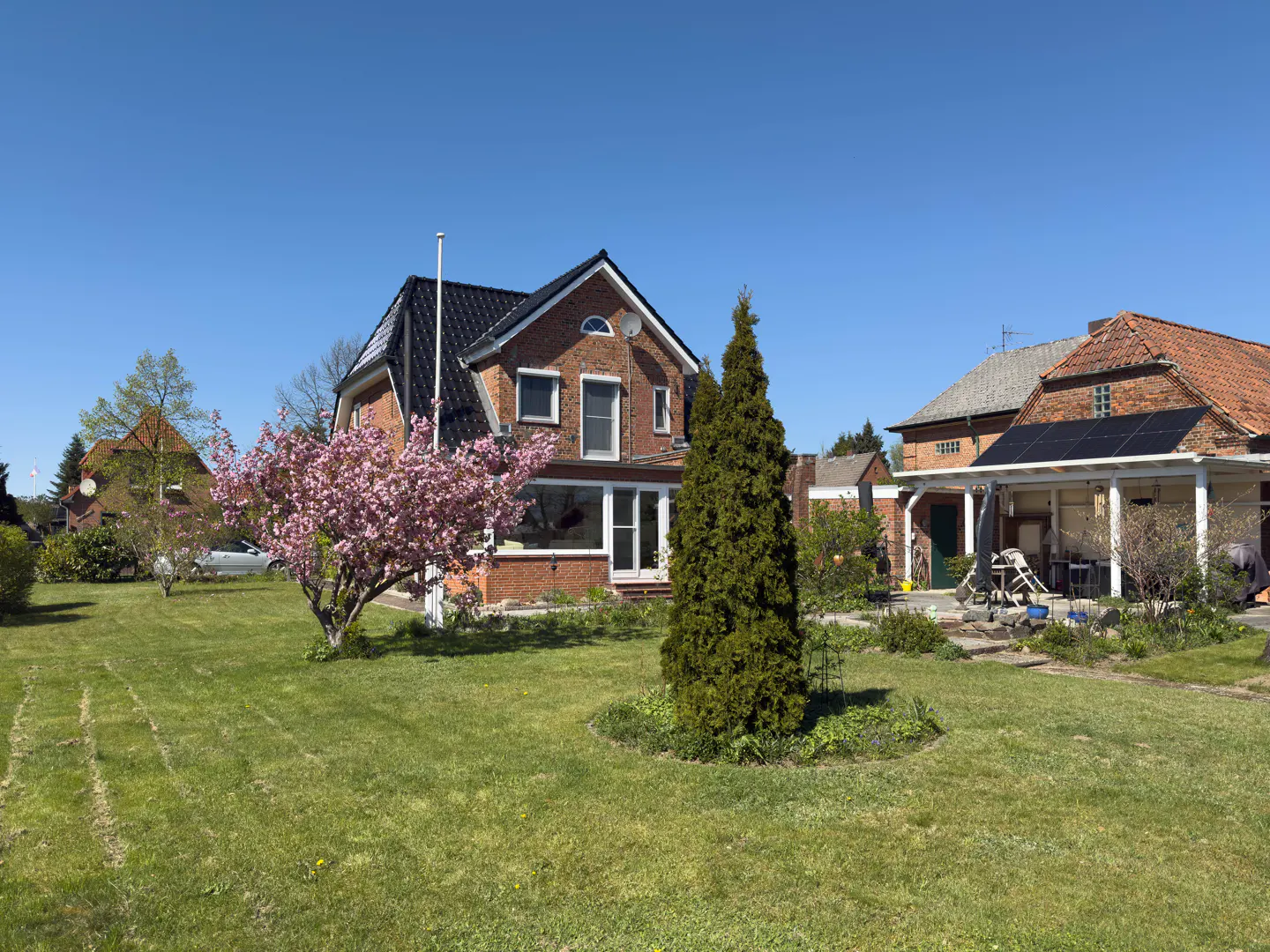 Exterior view of a red brick house with a black roof, surrounded by a green lawn, trees, and a pink flowering tree under a clear blue sky.