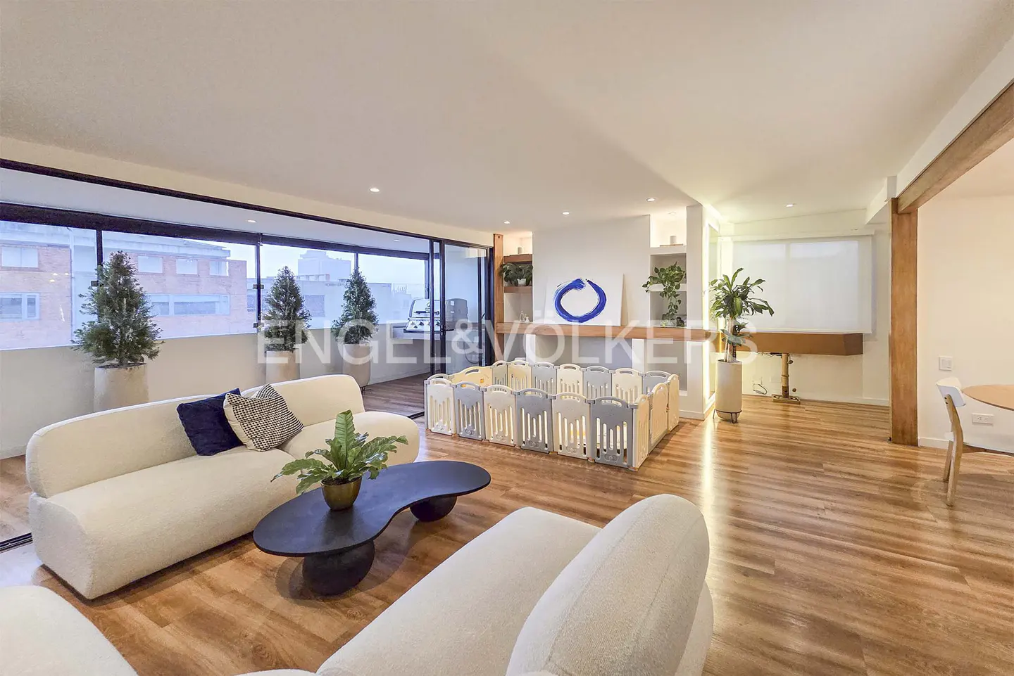 Bright living room with wood floors, white sofas, and a baby playpen. Large windows overlook a city. A black coffee table sits in the center.