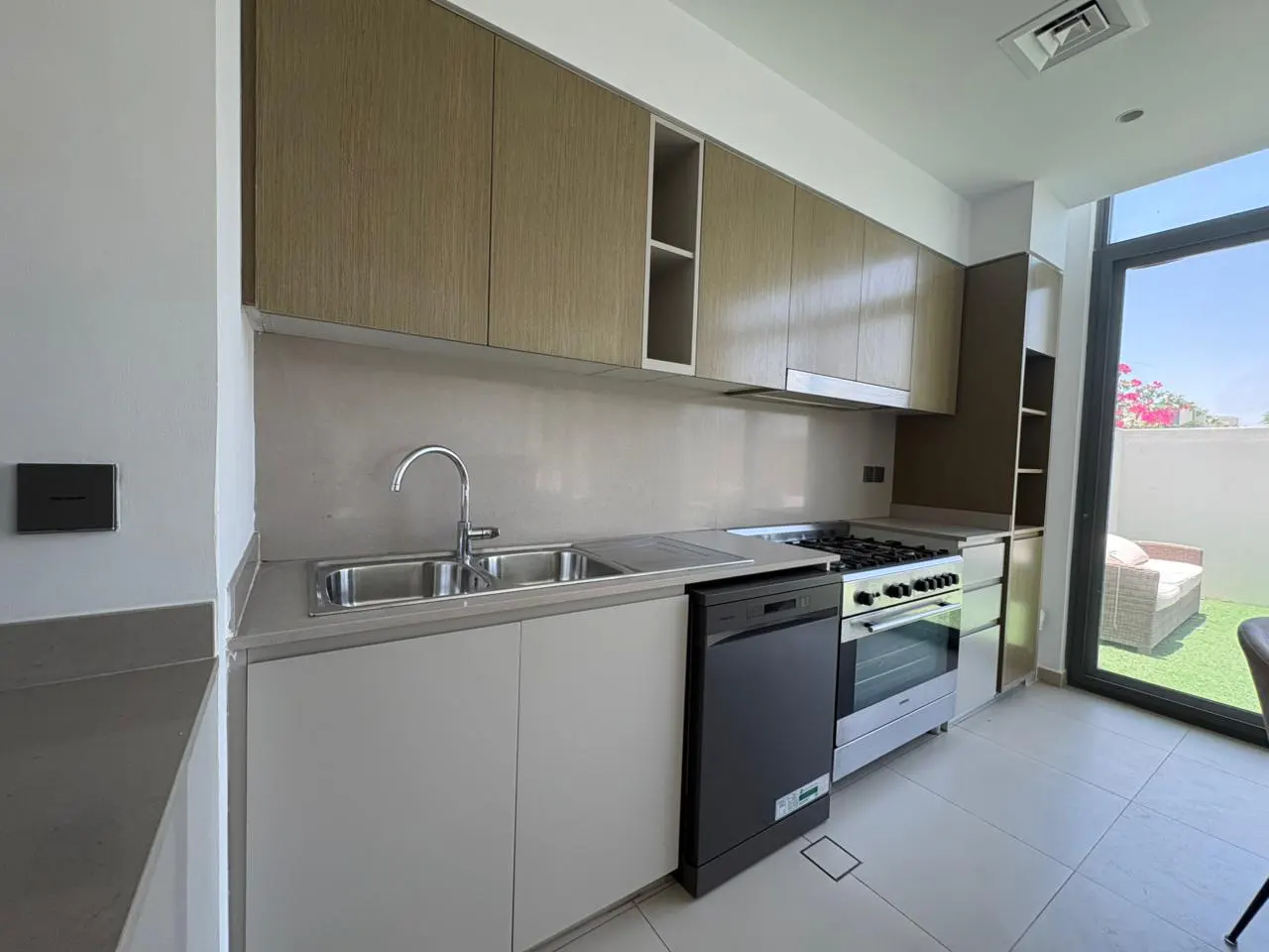 A modern kitchen with light wood cabinets, stainless steel sink, black dishwasher, and gas range. A patio is visible through a glass door.