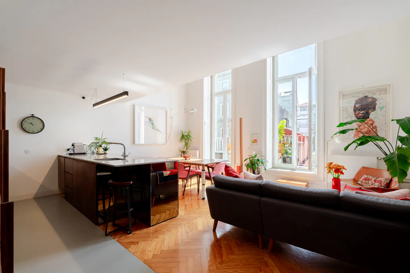Bright, open-concept apartment with a dark kitchen island, herringbone wood floors, and large windows. A black sofa faces the windows.