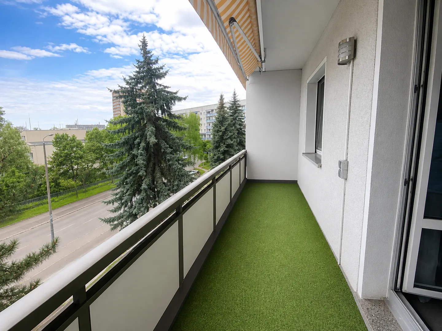 Apartment balcony with green turf, white walls, and a metal railing overlooking trees and a street.