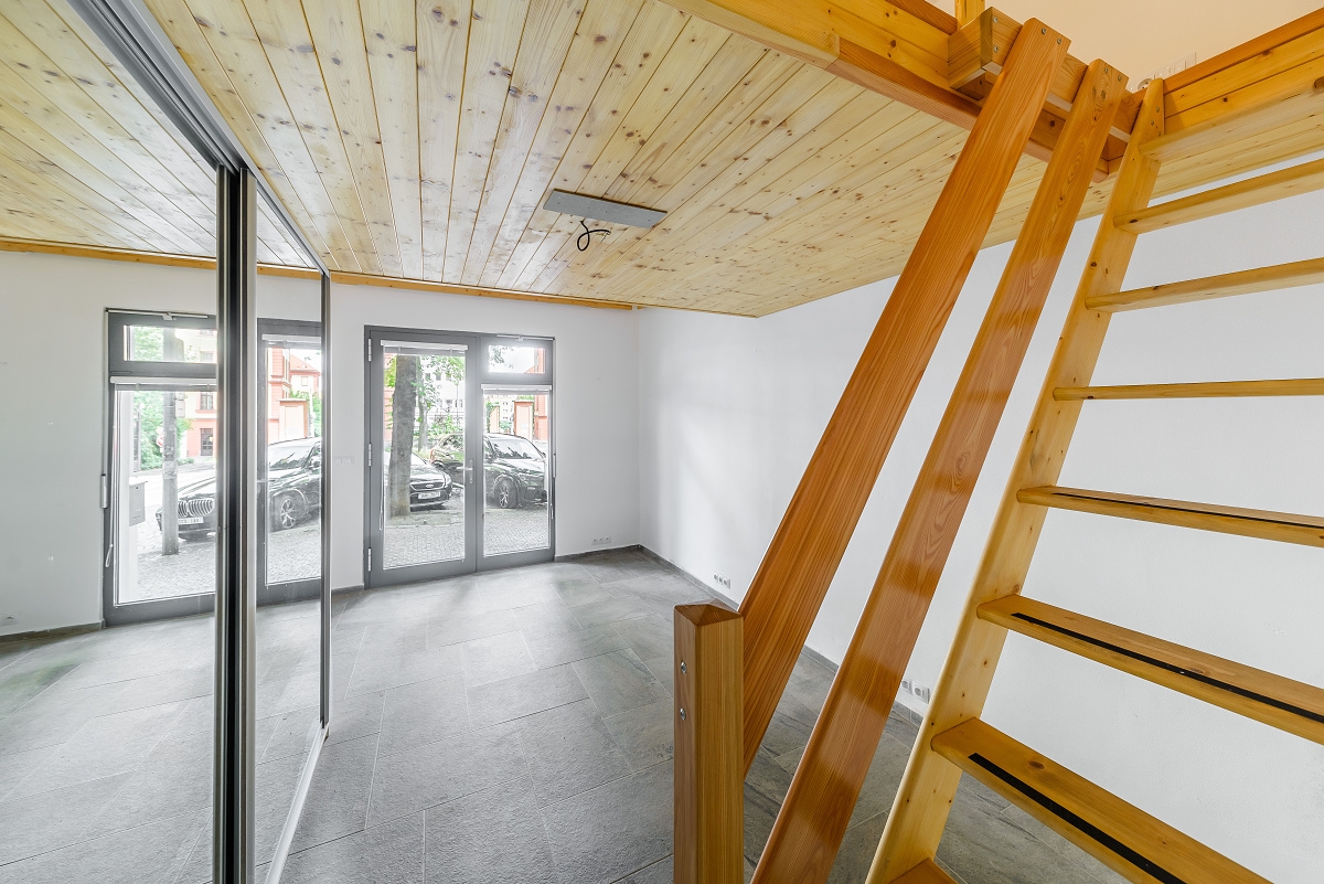 Modern room with grey tile floors, wooden ceiling, and a wooden staircase leading to a loft, with glass doors to a street. [AI-generated text]