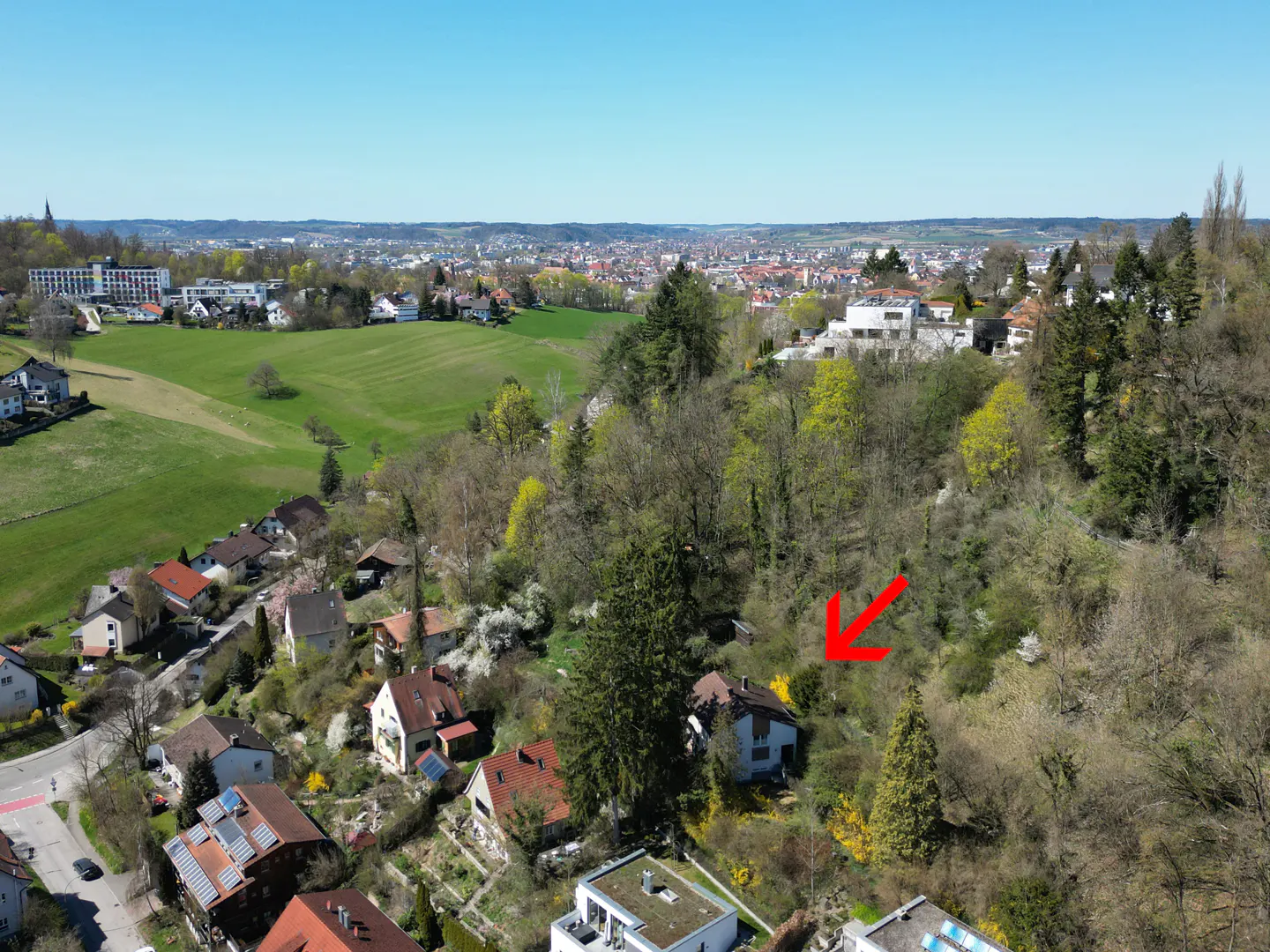 Aerial view of a white house with a brown roof, indicated by a red arrow, nestled among trees on a hillside. City in the background.