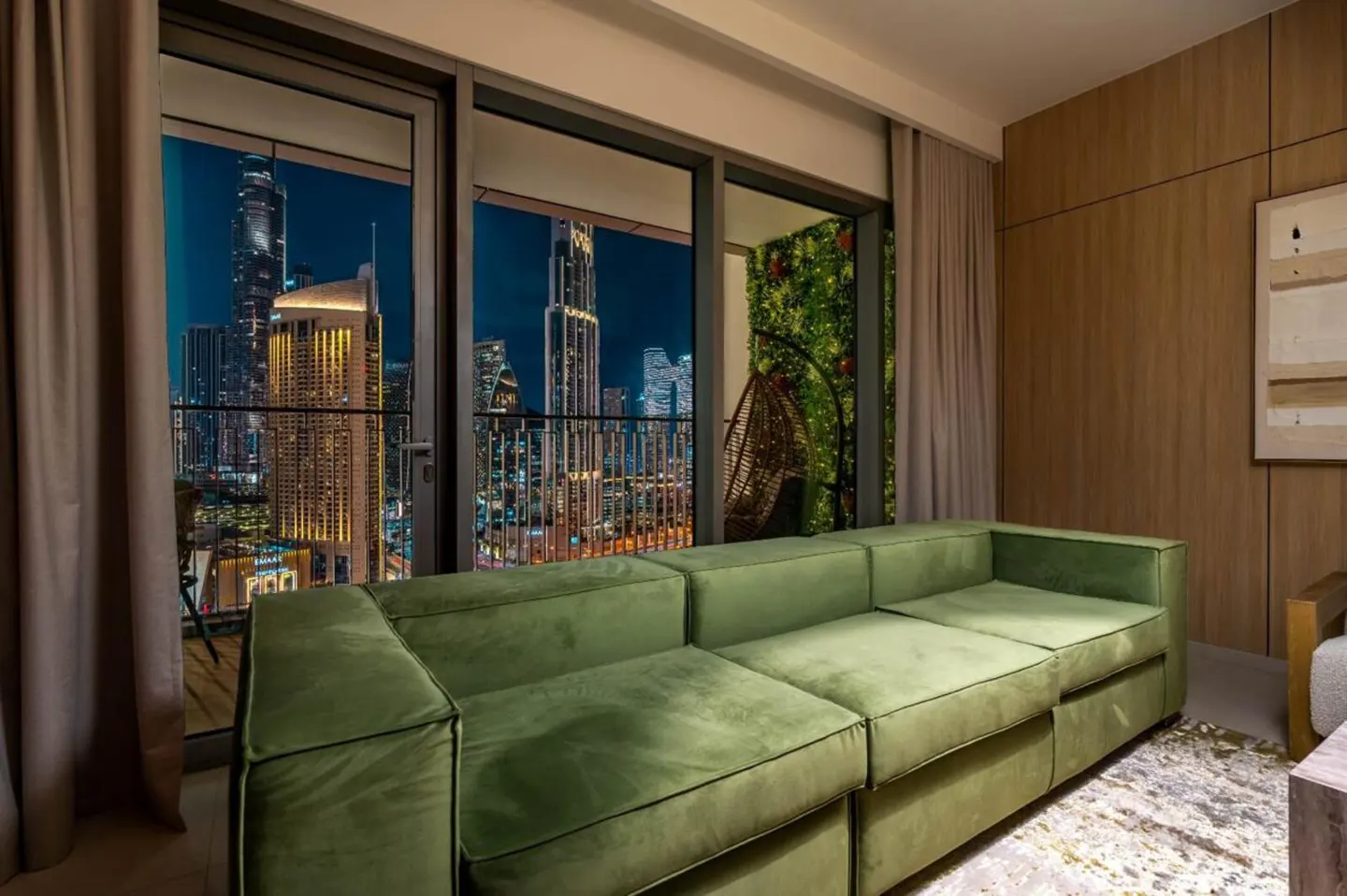 Living room with a green sofa and a balcony overlooking a city skyline at night. A hanging chair is on the balcony.