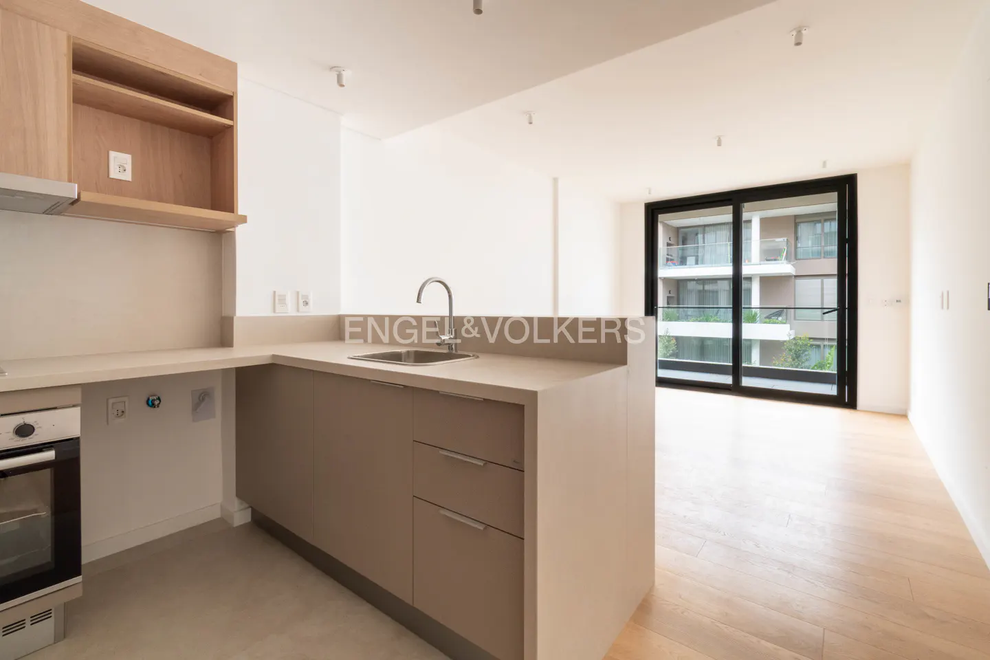 Modern kitchen with light wood cabinets, beige countertops, and a sink, opening to a bright living area with a balcony.