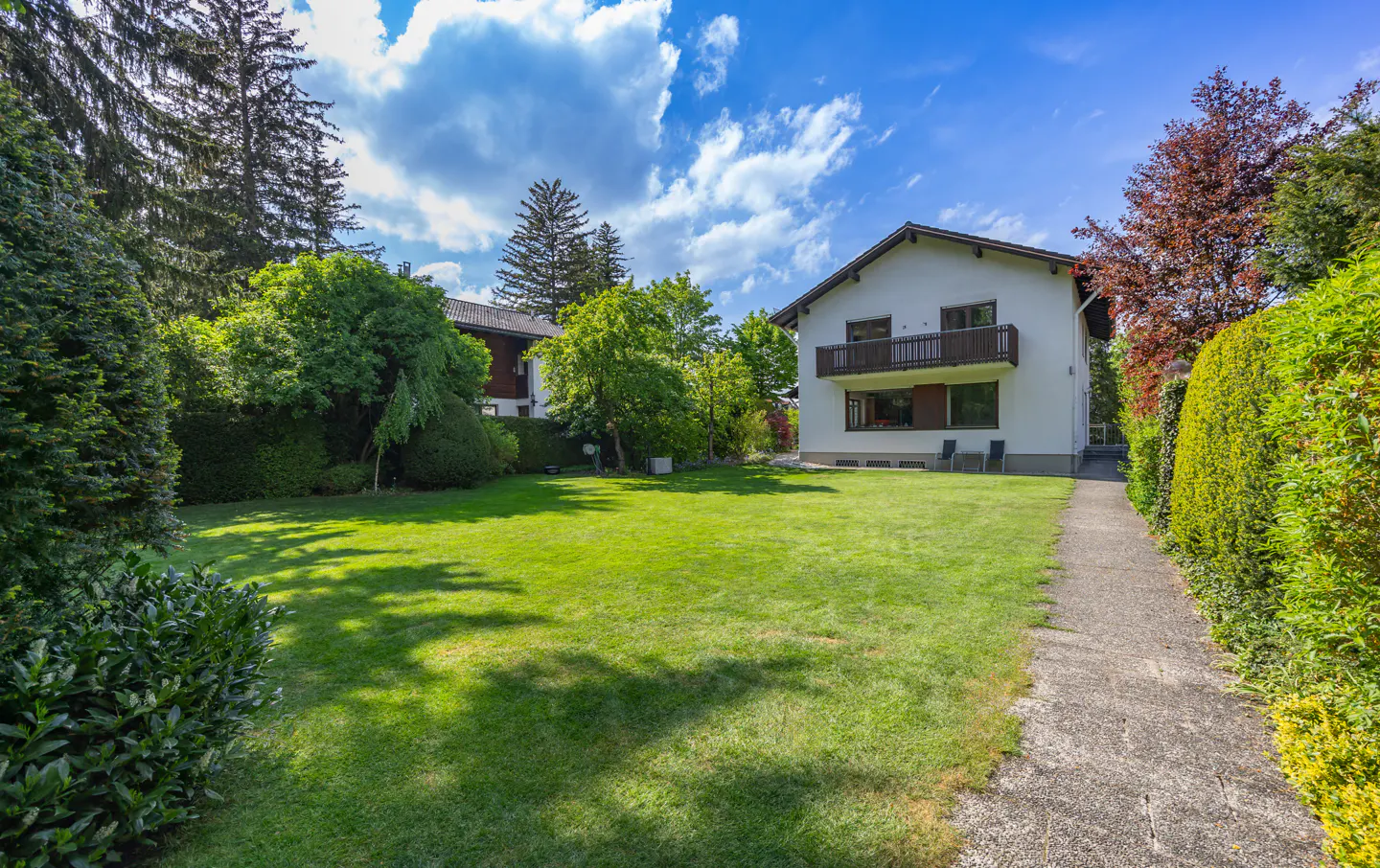 A white two-story house with a balcony overlooks a large green lawn, surrounded by trees and a stone pathway.