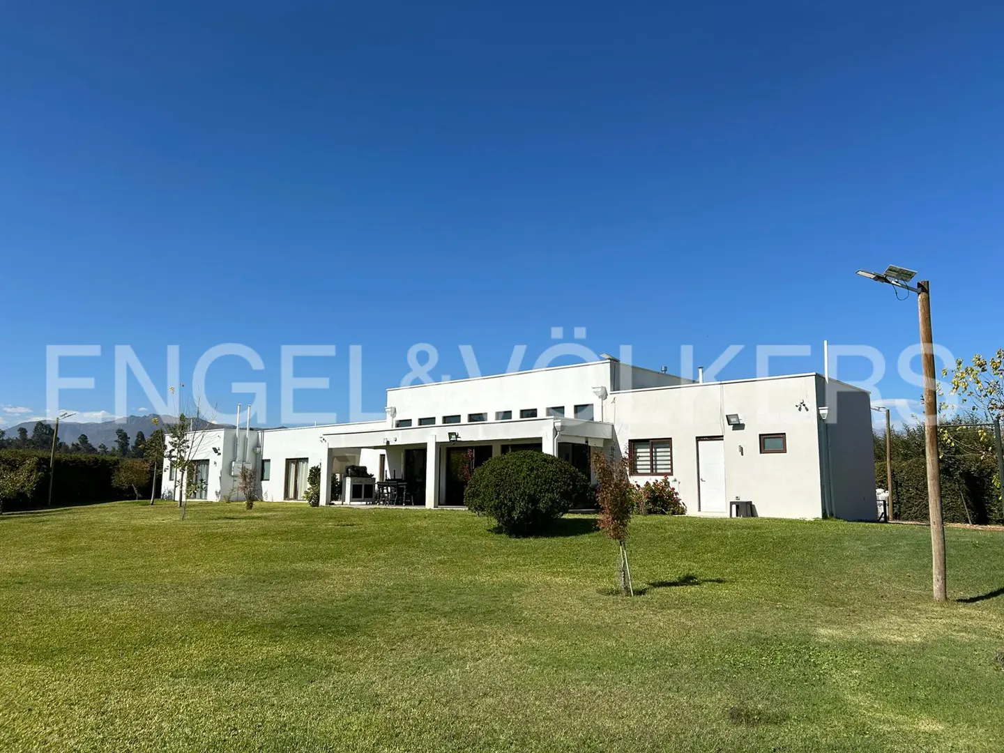 A modern, white, single-story house with a green lawn under a clear blue sky. The Engel & Völkers logo is visible.