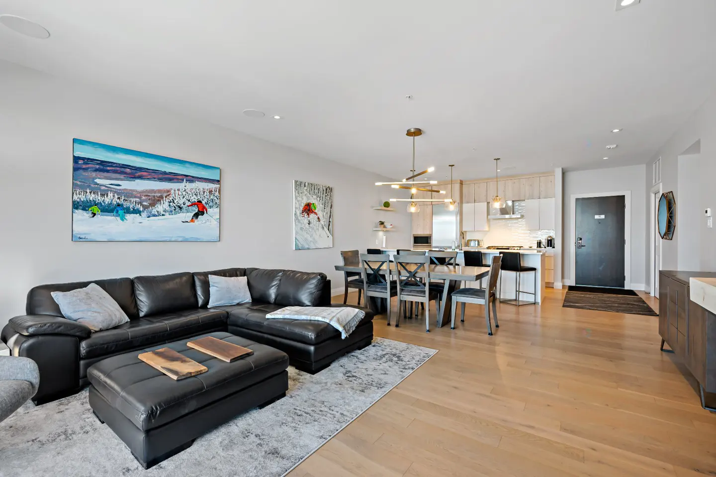 Open-concept living space with a black leather sectional, dining table, and kitchen. Skiing artwork adorns the walls.