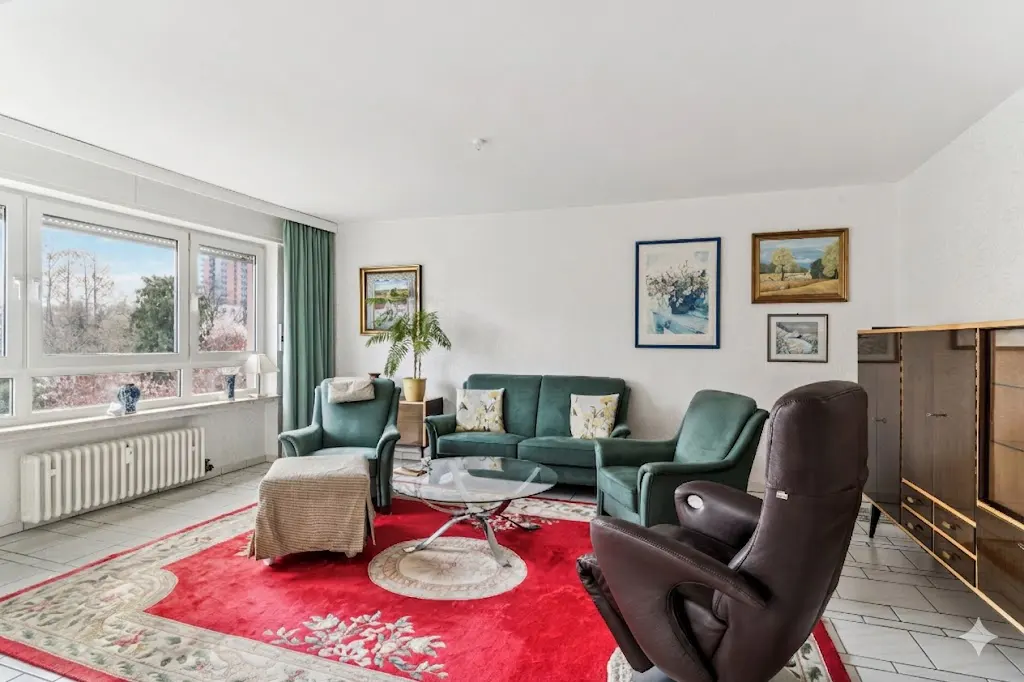 Bright living room with green sofa and armchairs, a red rug, and a large window with a view of trees.