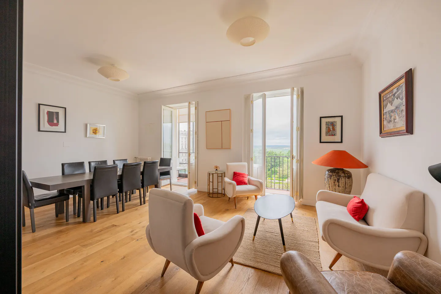 Bright living and dining area with hardwood floors, white walls, and neutral furniture with red accents. Balcony doors open to a green view.