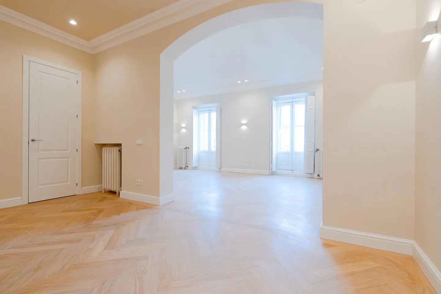 Bright, empty room with herringbone wood floors, beige walls, and white trim. An arched doorway leads to a sunlit room with two windows.