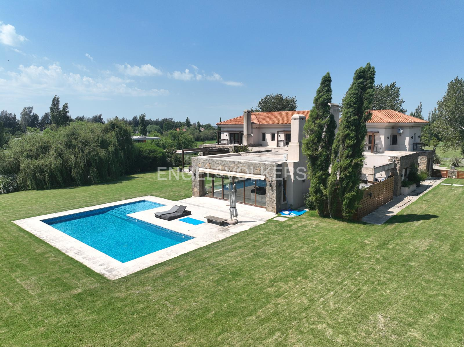 Aerial view of a luxury home with a pool, green lawn, and trees under a blue sky.