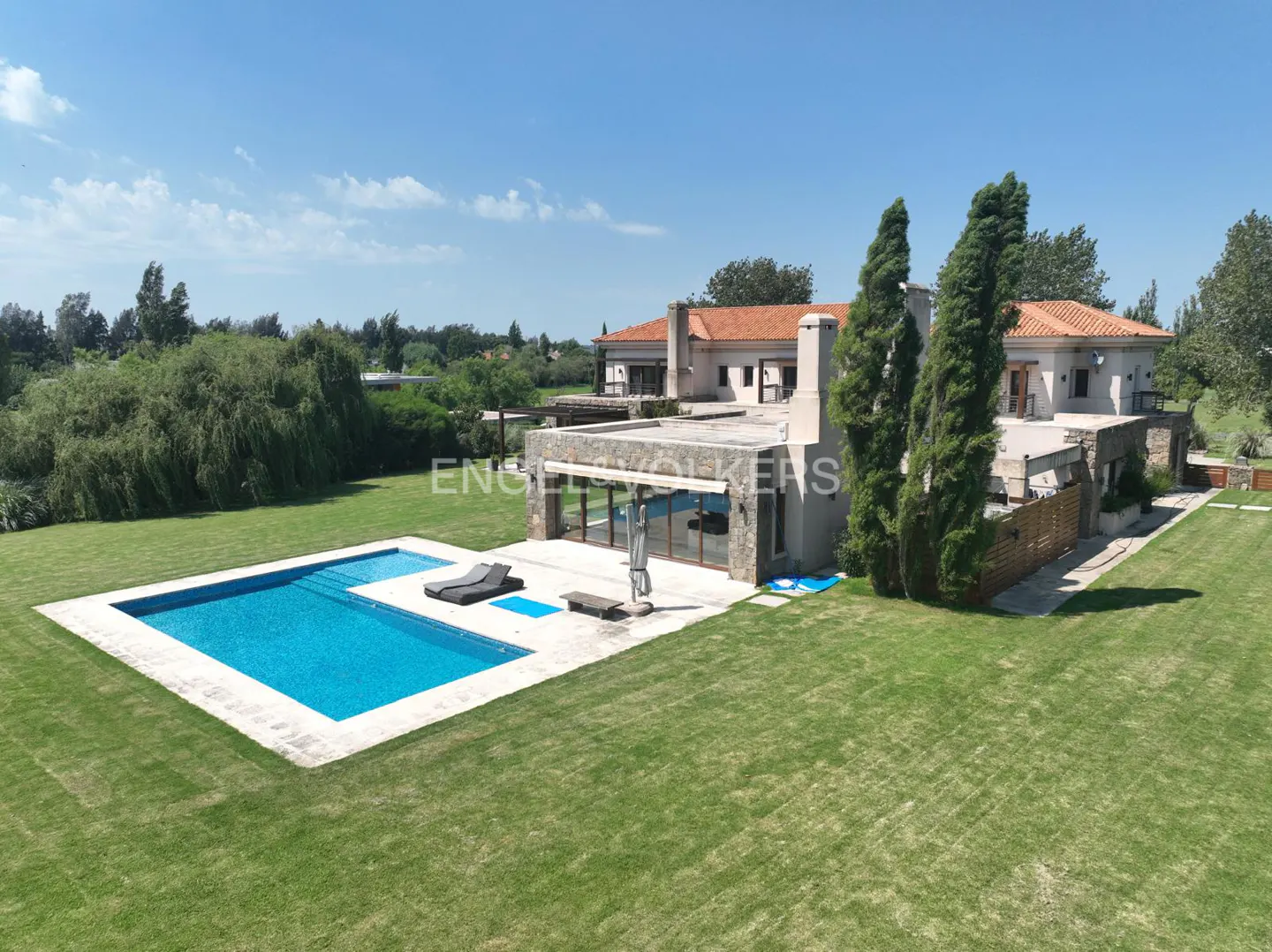 Aerial view of a luxury home with a pool, green lawn, and trees under a blue sky.