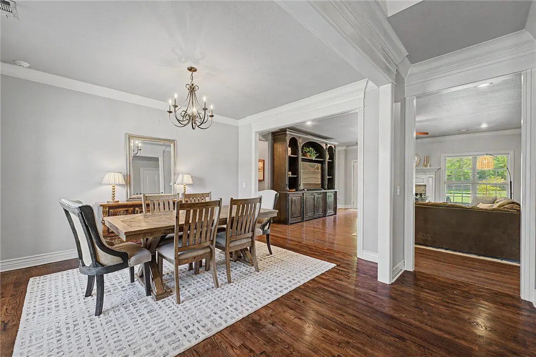 A dining room with a wooden table, chairs, and chandelier. A white rug sits under the table on a dark wood floor. A living room is visible through an open doorway.
