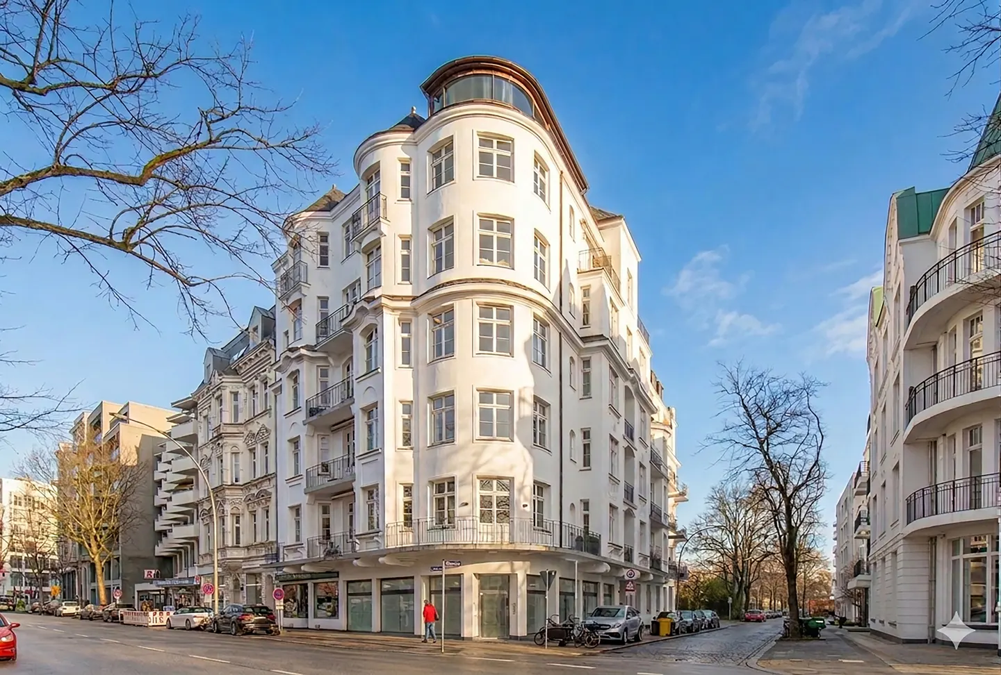 Street view of a multi-story white apartment building with balconies, cars parked along the street, and a blue sky.