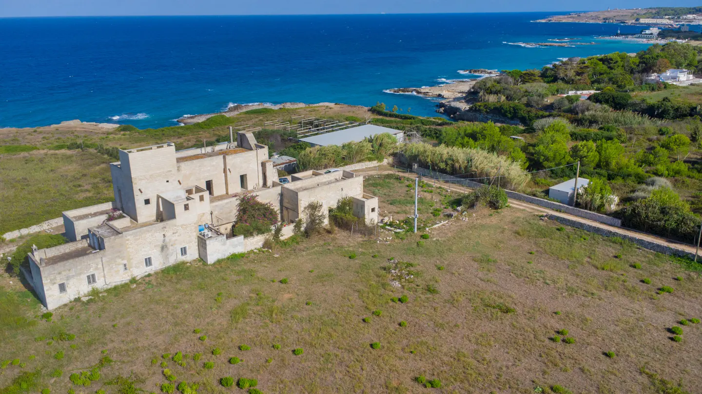Aerial view of a beige stone building with a tower, near the coast with blue water and green trees.