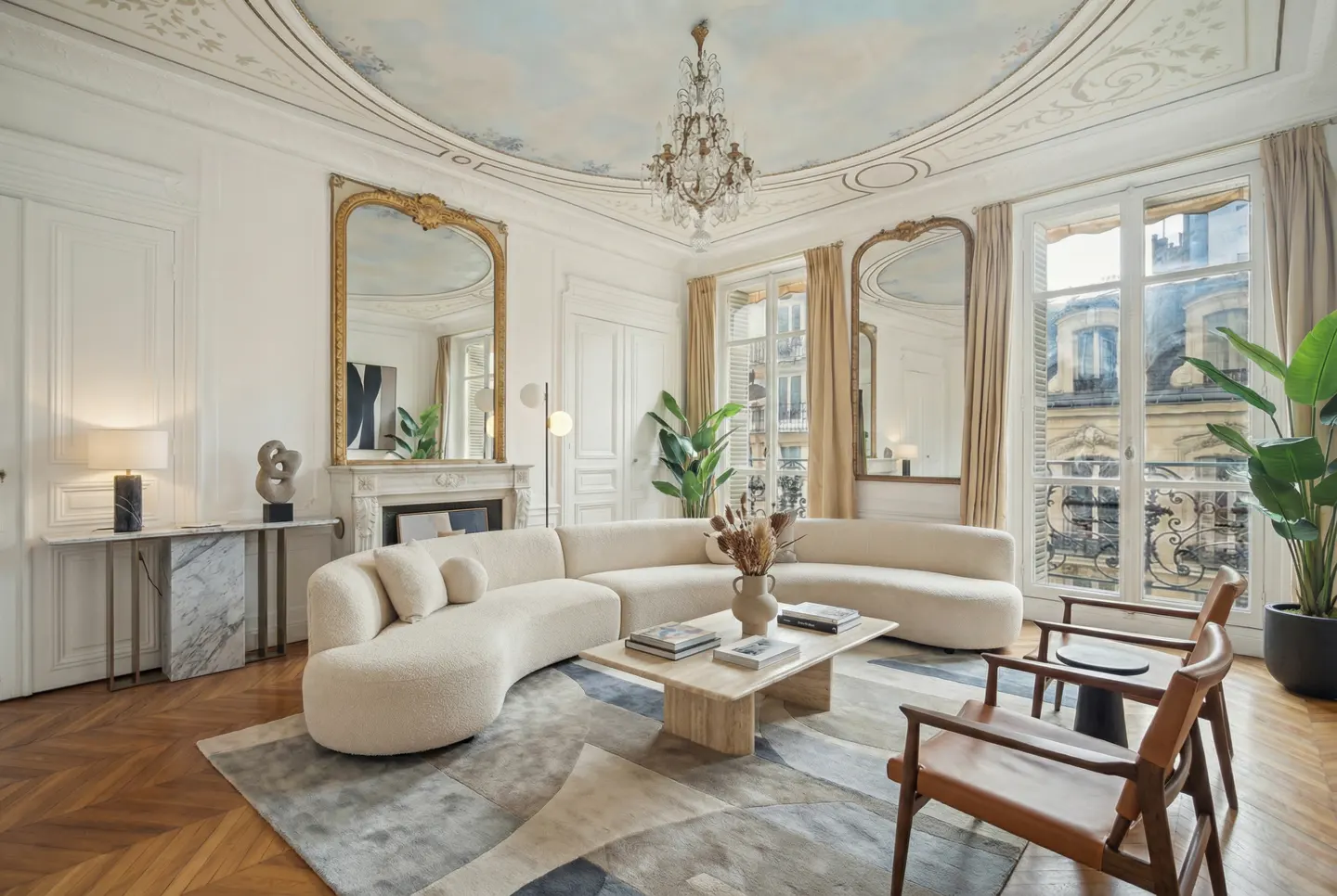 Elegant living room with a curved cream sofa, marble coffee table, crystal chandelier, and ornate mirrors on white walls.