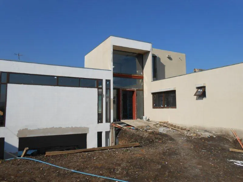 Modern, two-story house under construction with white and beige exterior walls, large windows, and a dirt yard.