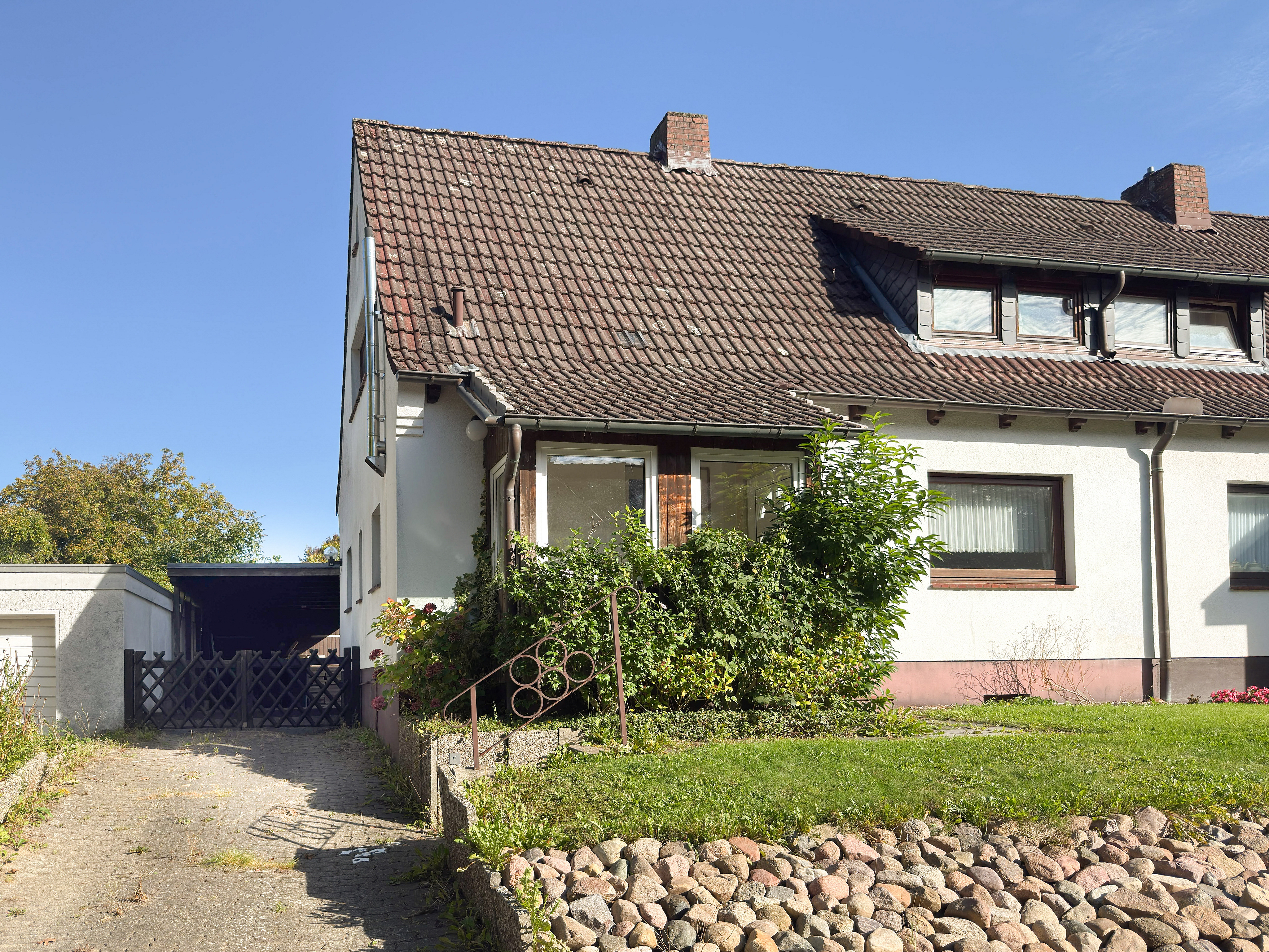 White semi-detached house with a brown tiled roof, a paved driveway, a small garden, and a stone retaining wall. [AI-generated text]
