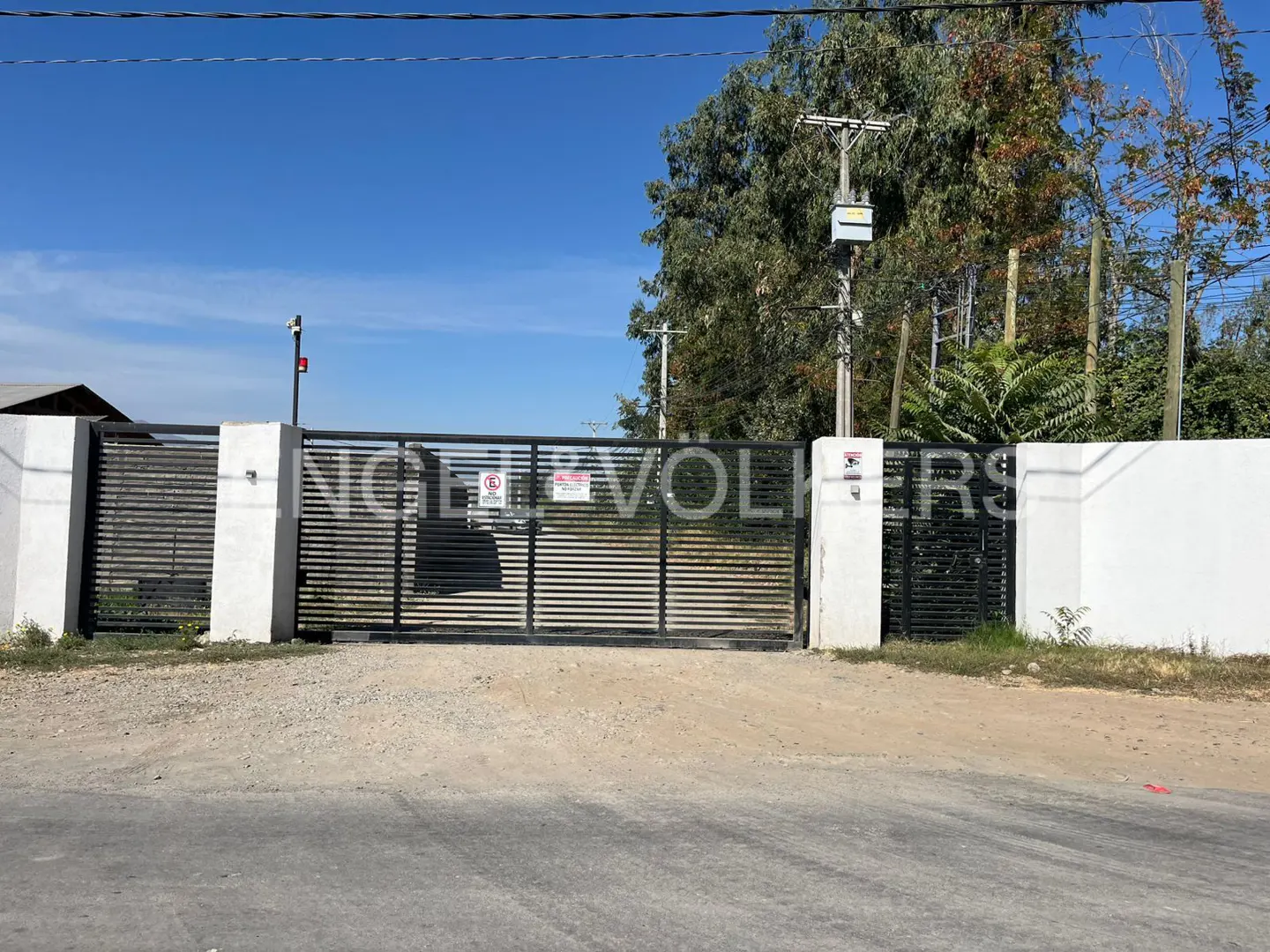 Black metal gate with white pillars, gravel driveway, and trees in the background under a blue sky.
