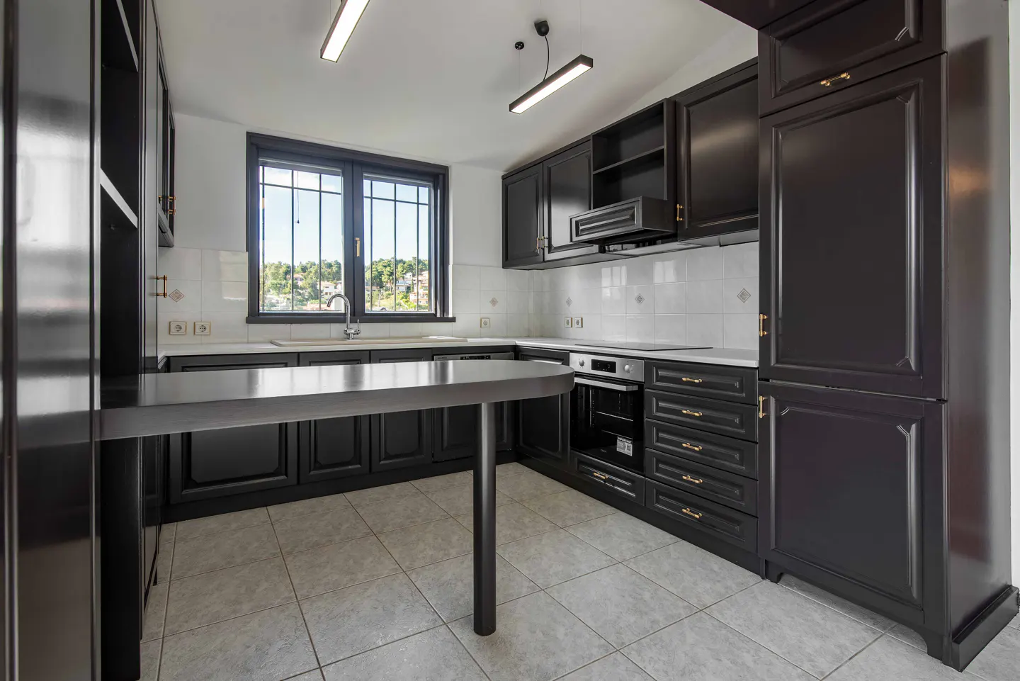 A modern kitchen with dark cabinets, white backsplash, and a gray tiled floor. A window overlooks a green landscape.