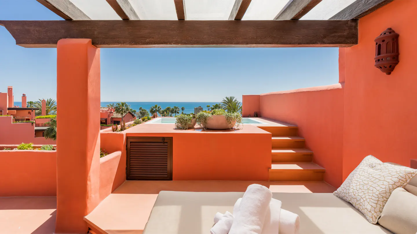 Terracotta patio with a daybed, white towels, and a view of the ocean and a rooftop pool.