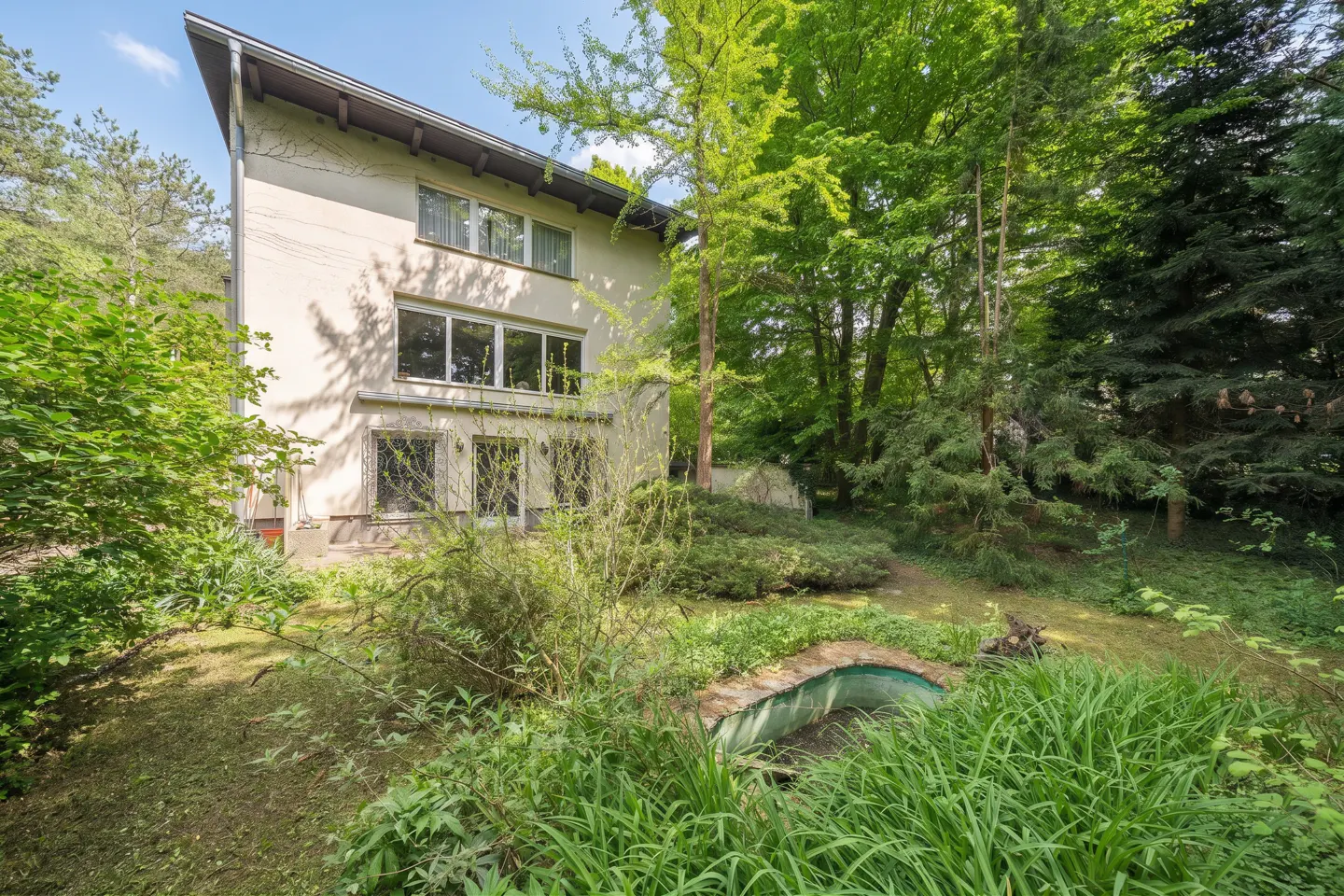 Two-story beige house with a sloped roof, surrounded by lush green trees and foliage. A small, arched bridge is in the foreground.