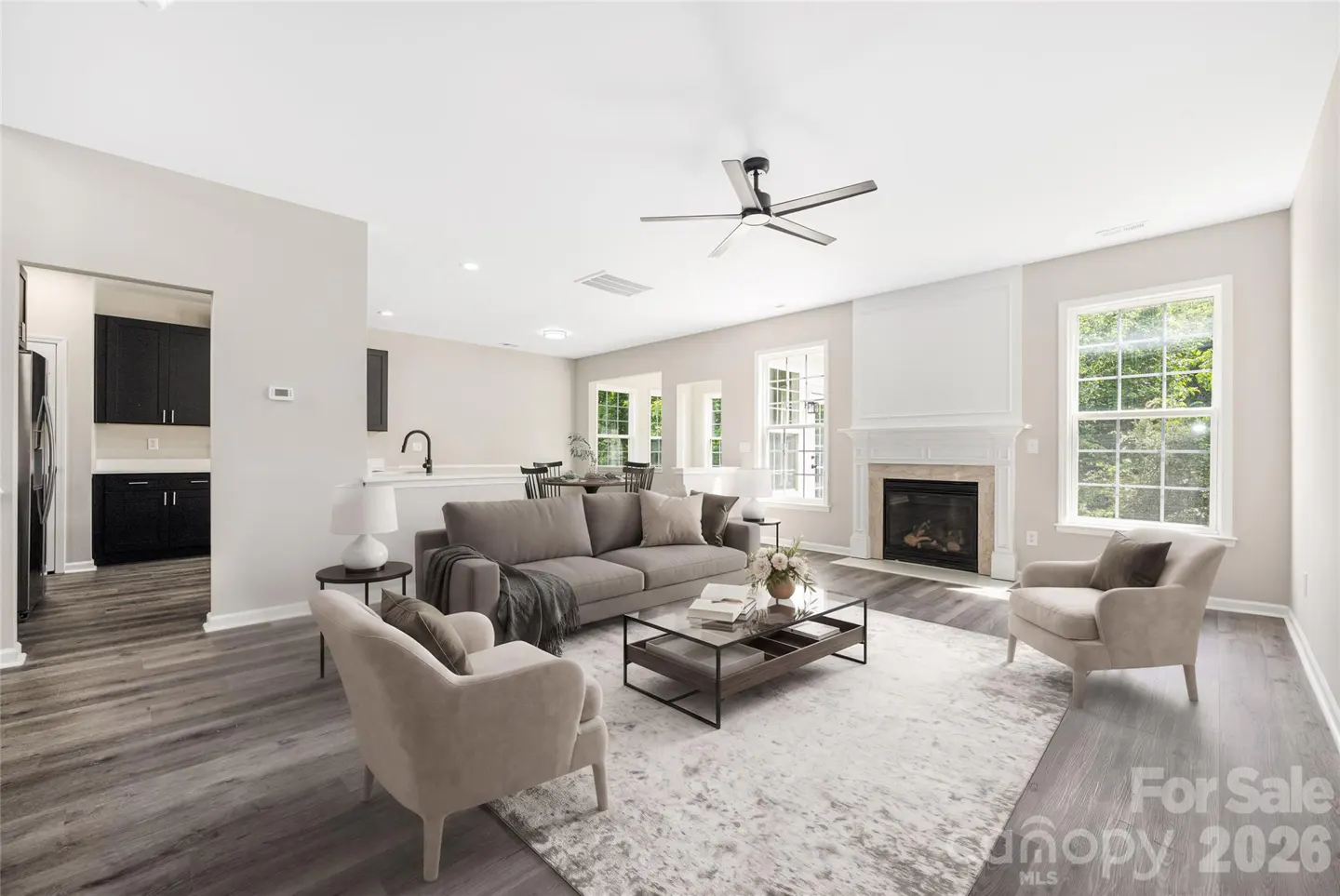 Open-concept living room with gray sofa, two beige armchairs, glass coffee table, fireplace, and view of dark kitchen.