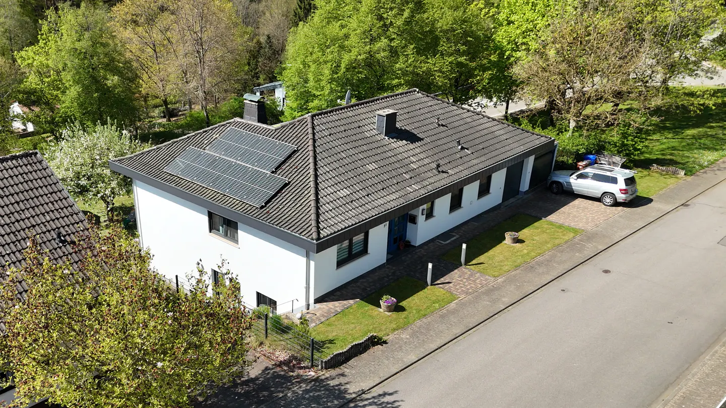 Aerial view of a white house with solar panels on a gray roof, a silver SUV parked in the driveway, and green trees in the background.