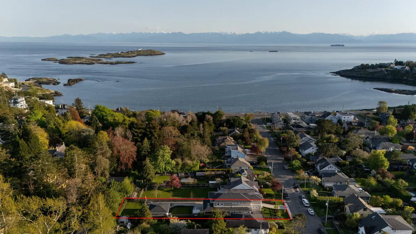 Aerial view of a gray house with a red border, trees, and ocean view. Mountains are visible in the distance.