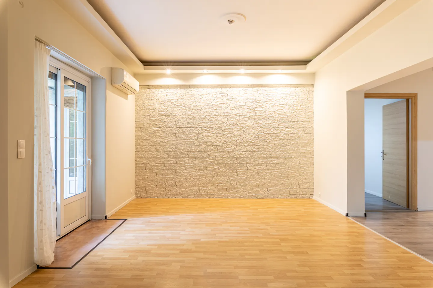 Bright, empty room with light wood floors, a stone accent wall, and a glass door with white curtains.