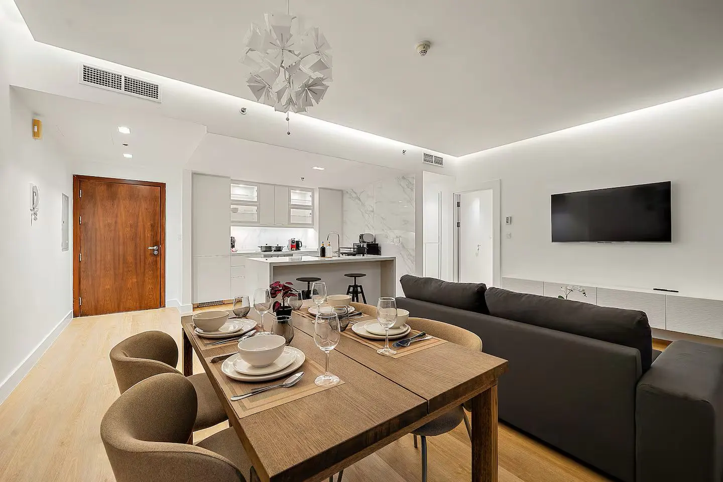 Open-concept living space with a wood dining table set for four, a gray sofa, and a white kitchen with marble backsplash.