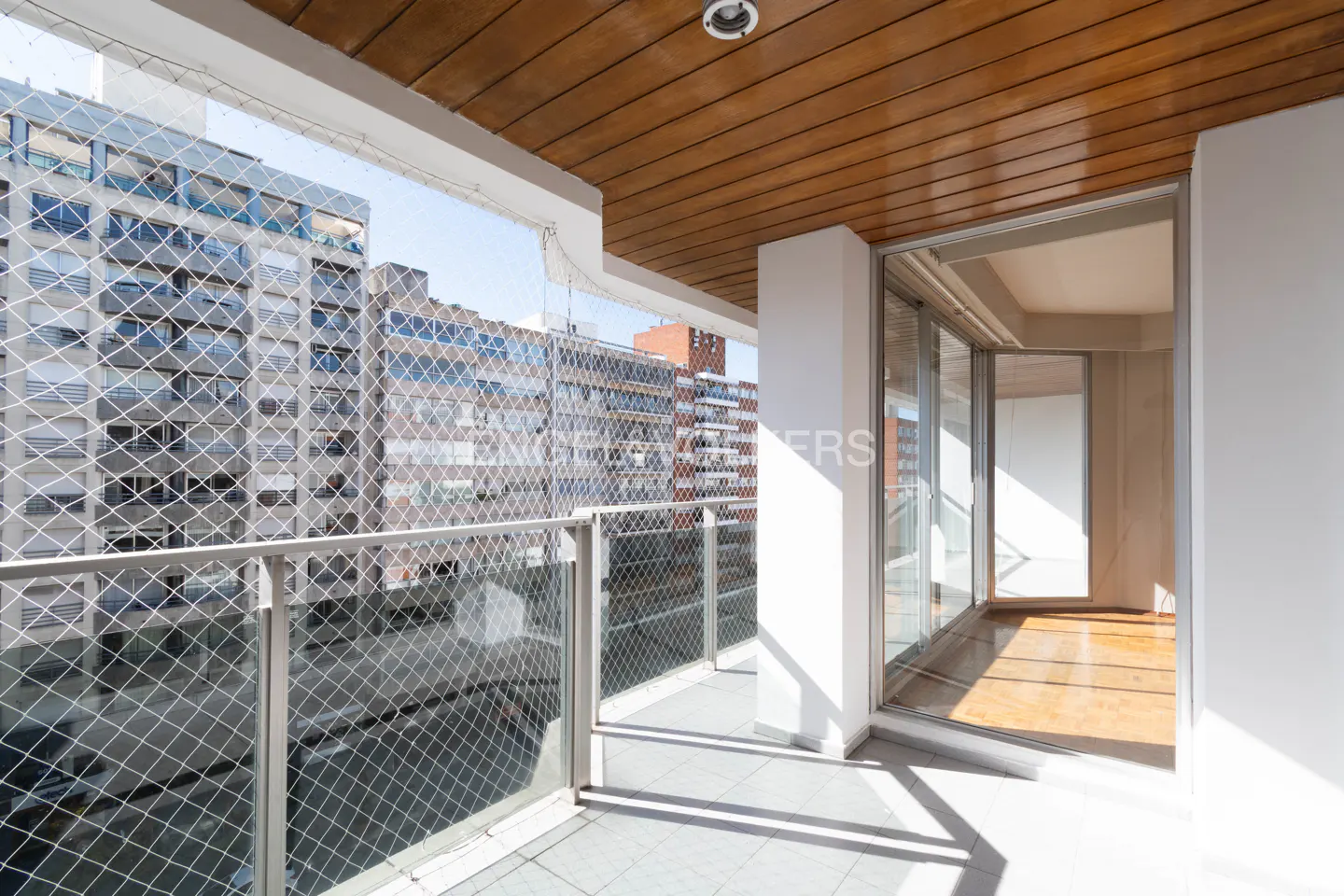 Balcony view with a safety net, gray tile floor, and a wood-paneled ceiling. Sliding glass doors lead to a room with hardwood floors. City buildings are visible in the background.