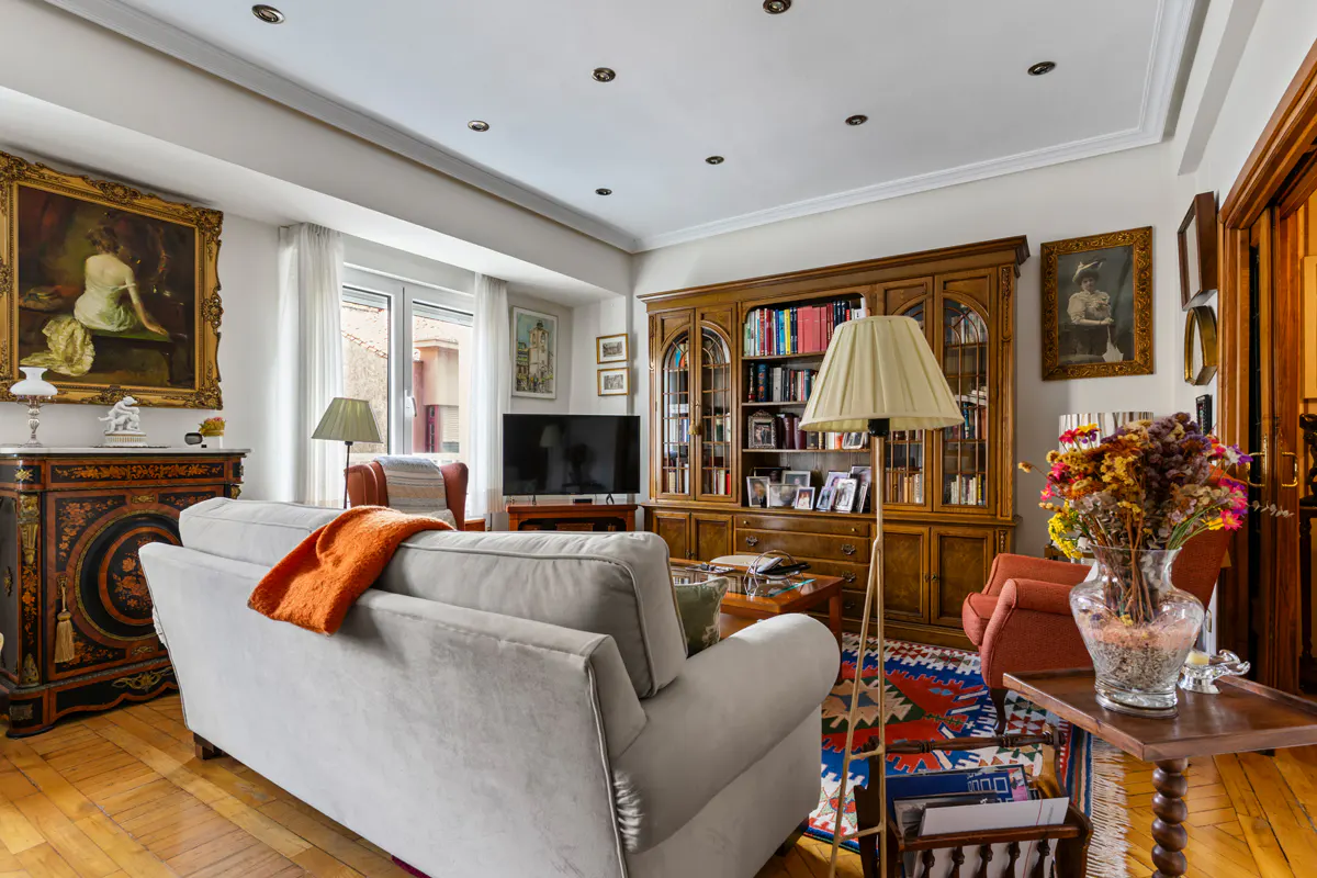 Classic living room with a light grey sofa, orange throw, wooden bookcase, patterned rug, and vintage decor on wood floors.