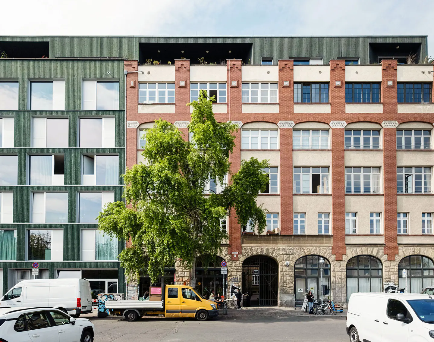 A street view of a modern green building next to a historic red brick building with a large tree in front.