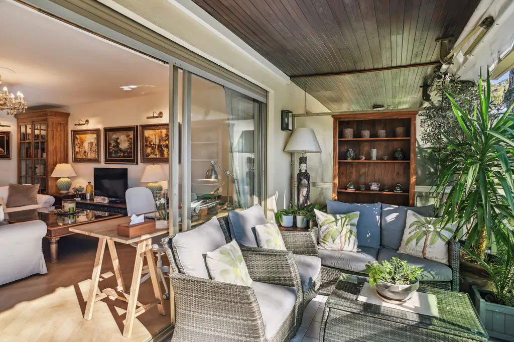 A sunlit patio with wicker furniture, blue cushions, and palm-leaf pillows, next to an open living room with a wooden cabinet and paintings.