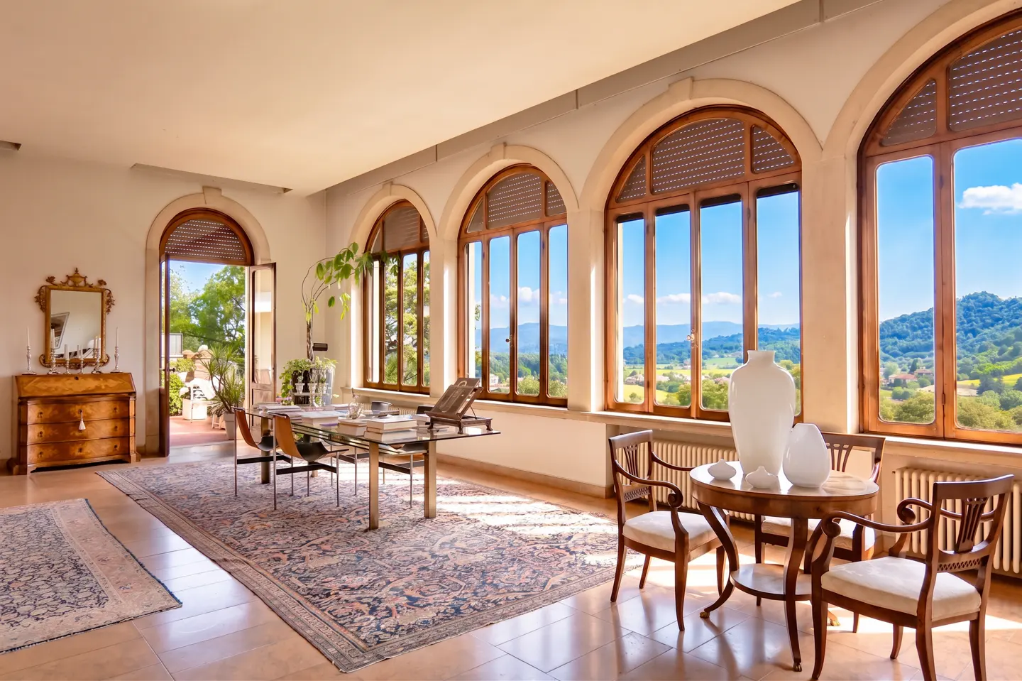 Bright sunroom with arched windows framing a green landscape. A glass table, chairs, and a round table with vases furnish the room.