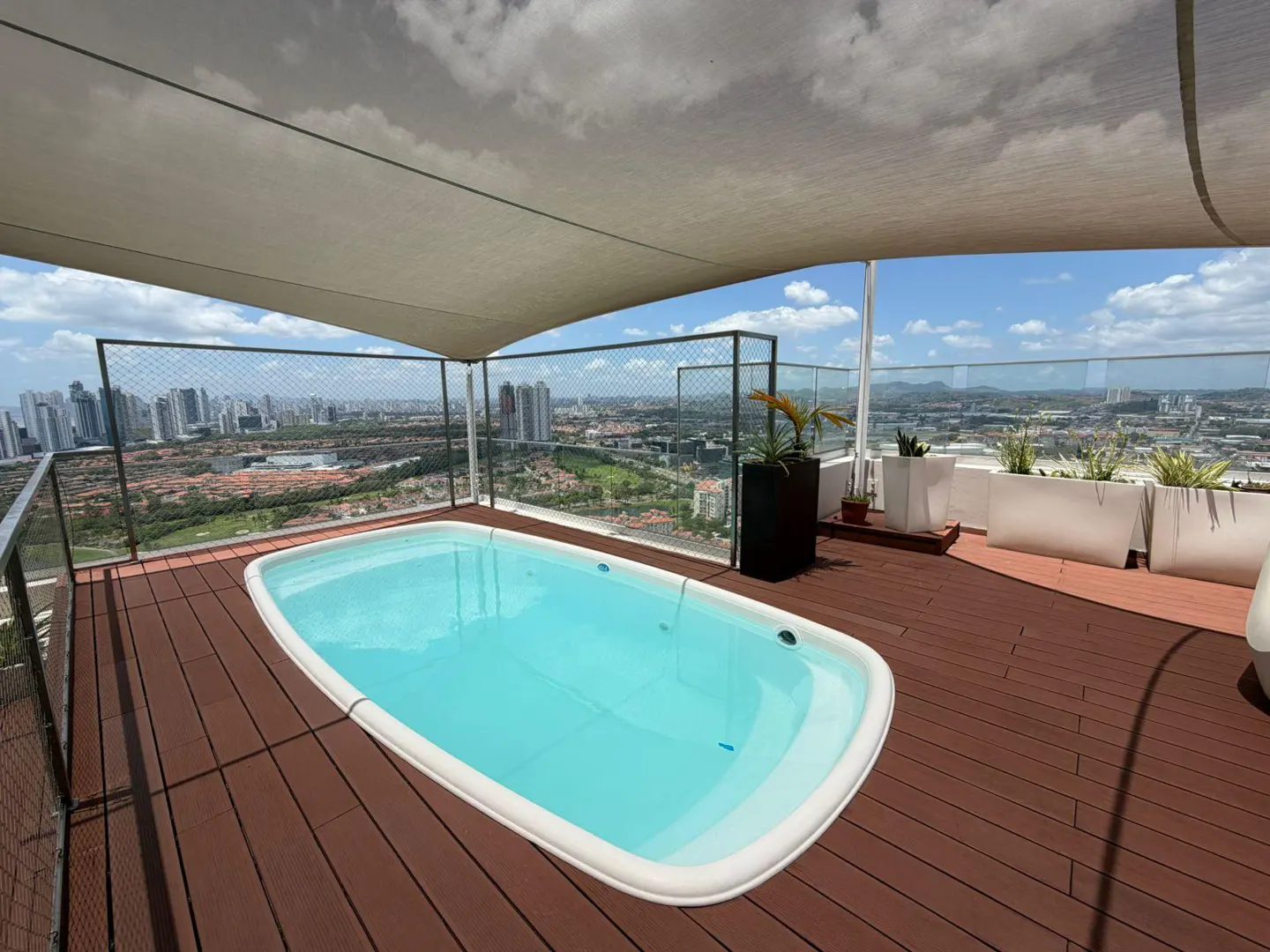 Rooftop pool with city view. A white pool filled with blue water sits on a brown deck, under a beige awning. Planters line the edge.