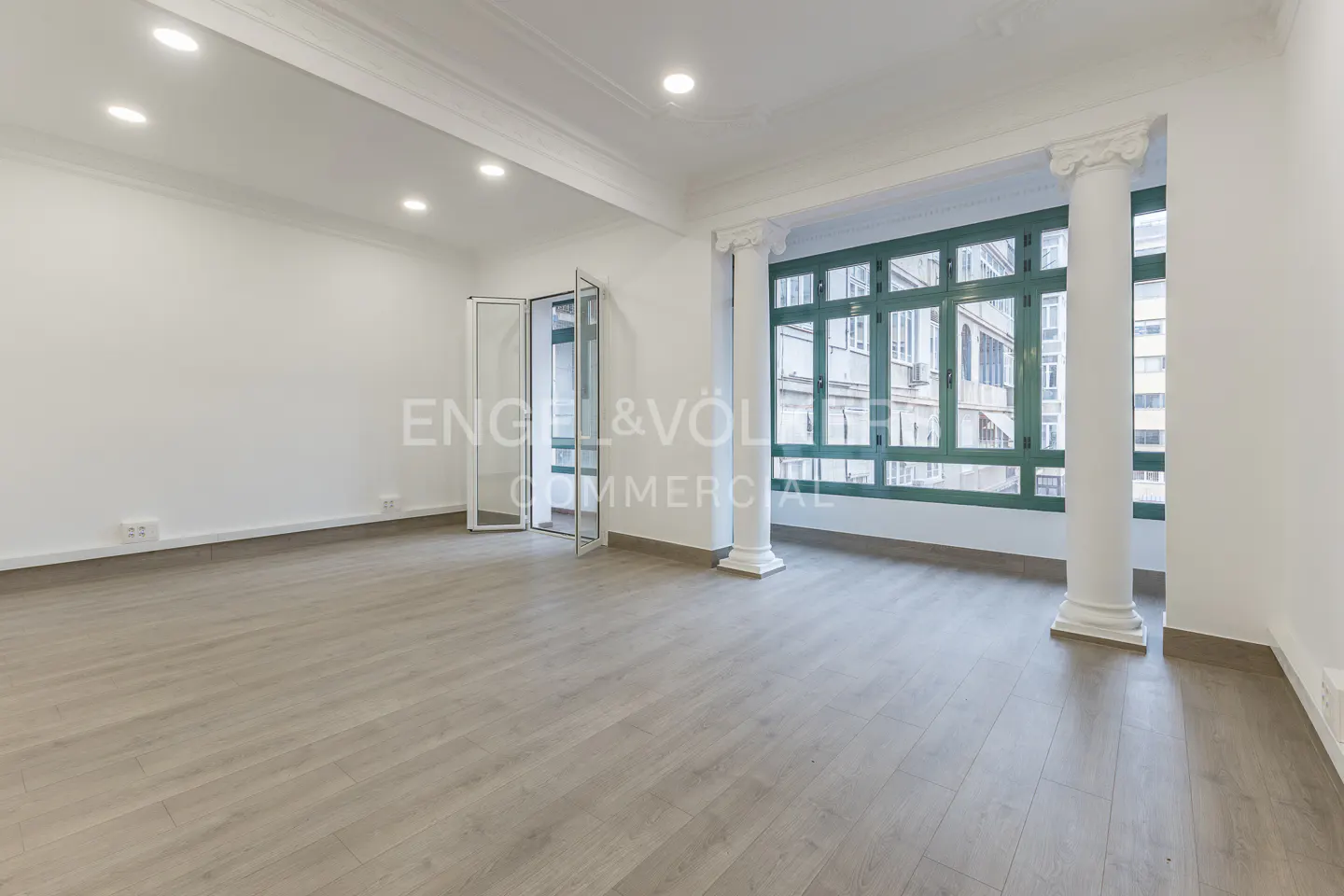 Bright, empty room with light wood floors, white walls, and large green-framed windows with white columns. Glass doors are open.