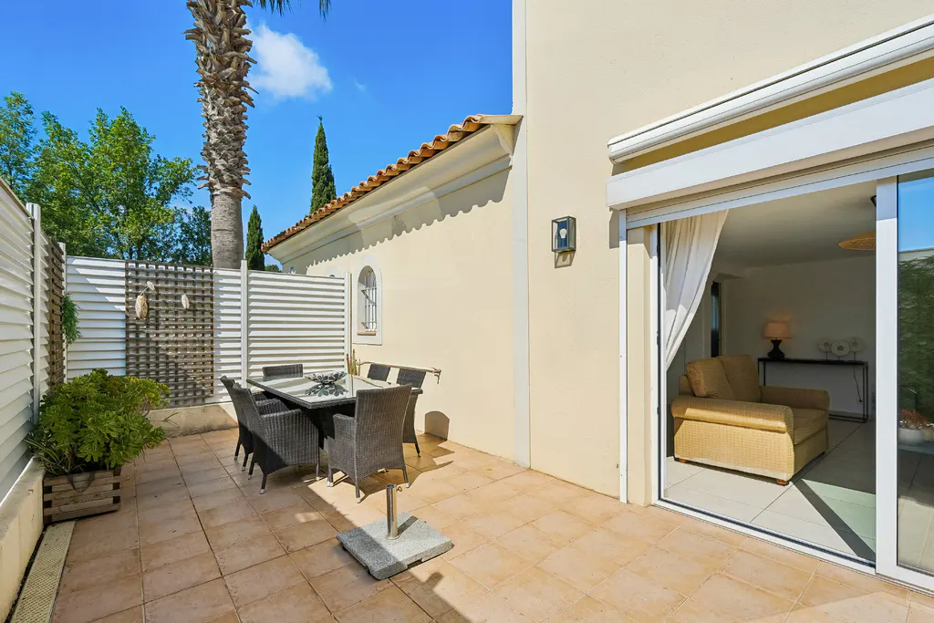 Outdoor patio with a table and chairs, a potted plant, and a sliding glass door leading to a living room with a yellow armchair.