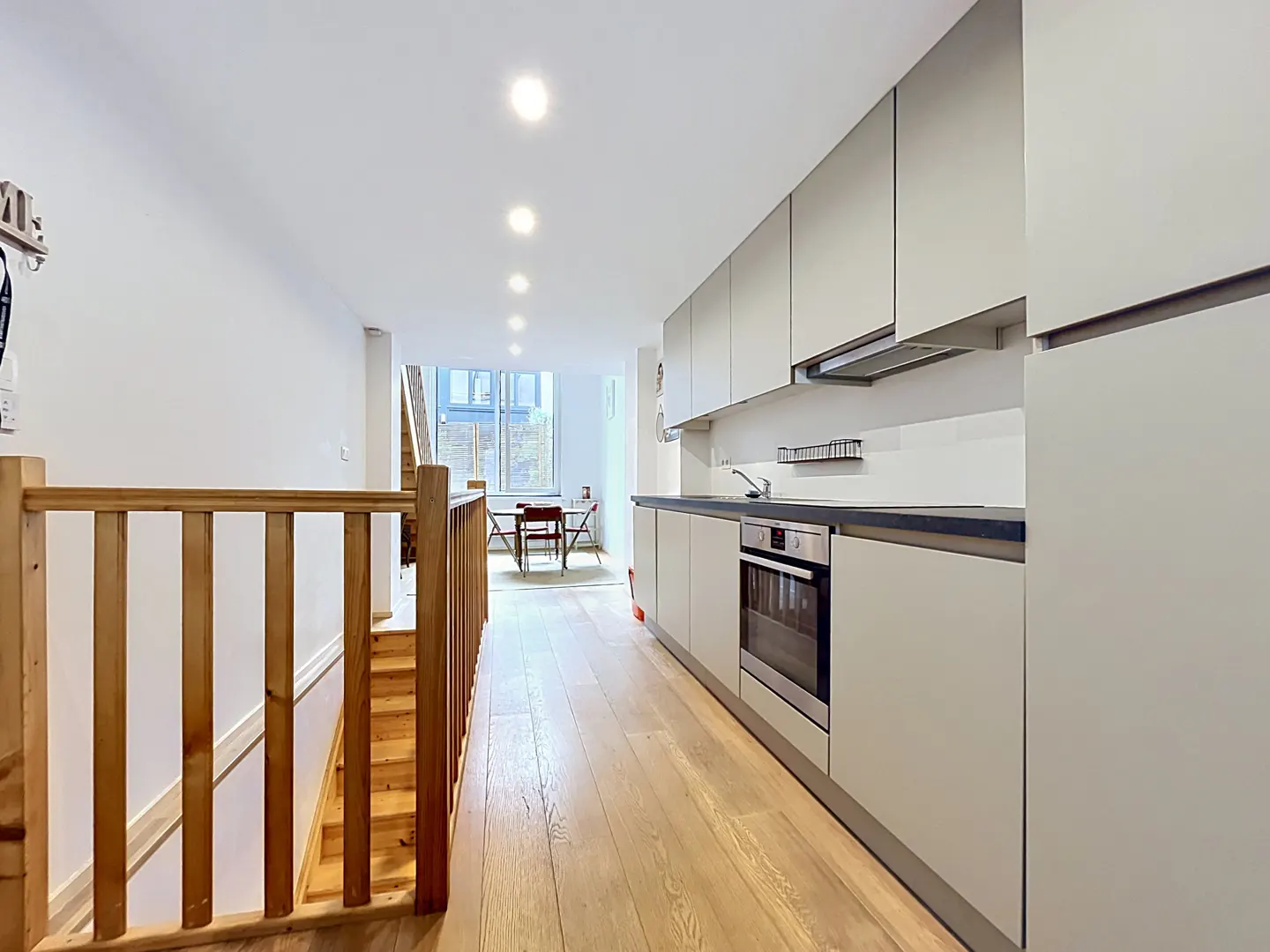 A long, narrow kitchen with light wood floors, gray cabinets, and a wooden staircase on the left. A table and chairs are visible in the background.